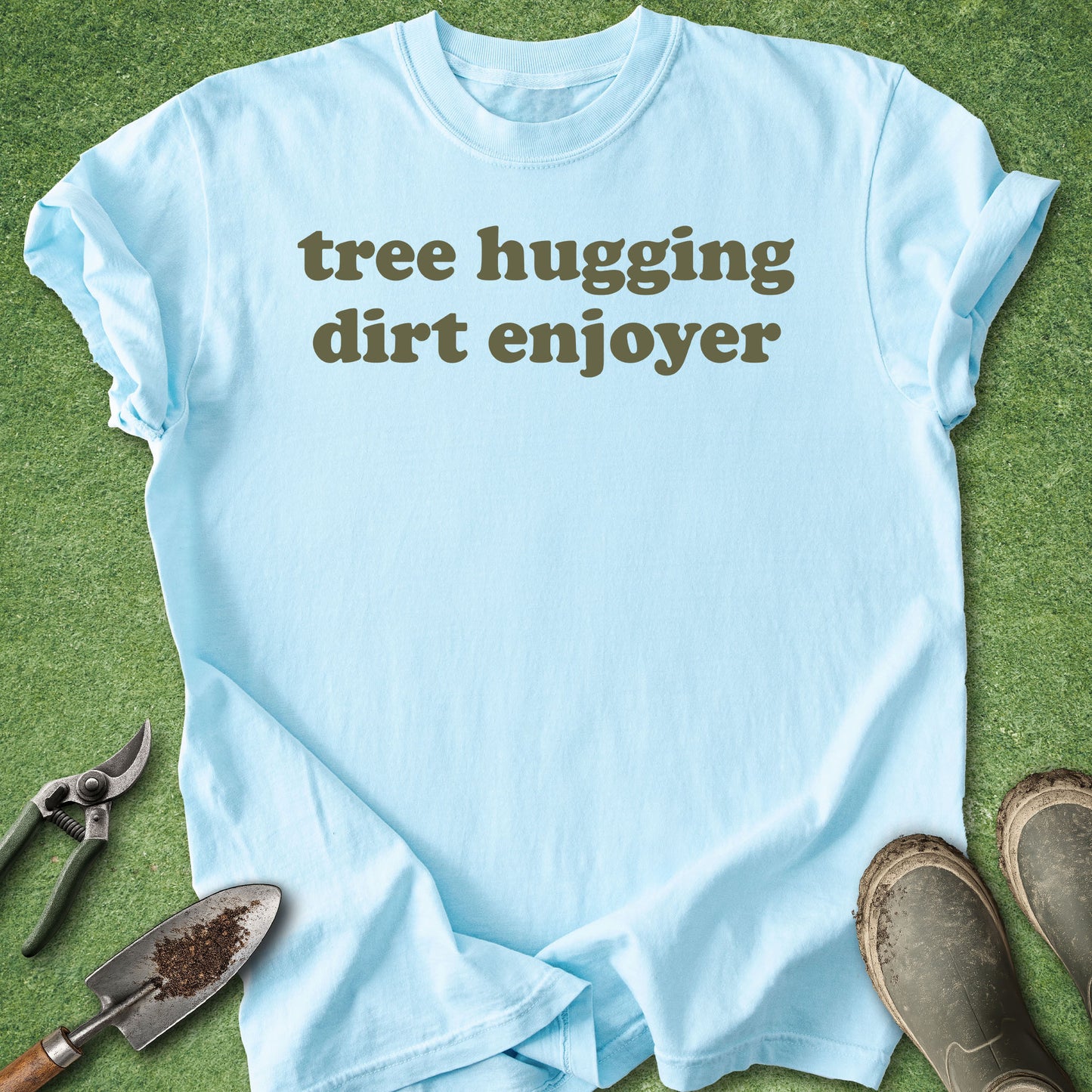 Light blue t-shirt with 'tree hugging dirt enjoyer' text on a grass background with gardening tools and boots.