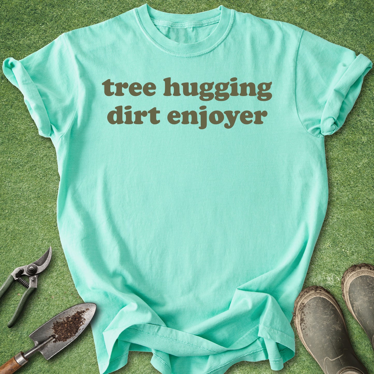 Mint green t-shirt with 'tree hugging dirt enjoyer' text on a grass background with gardening tools.