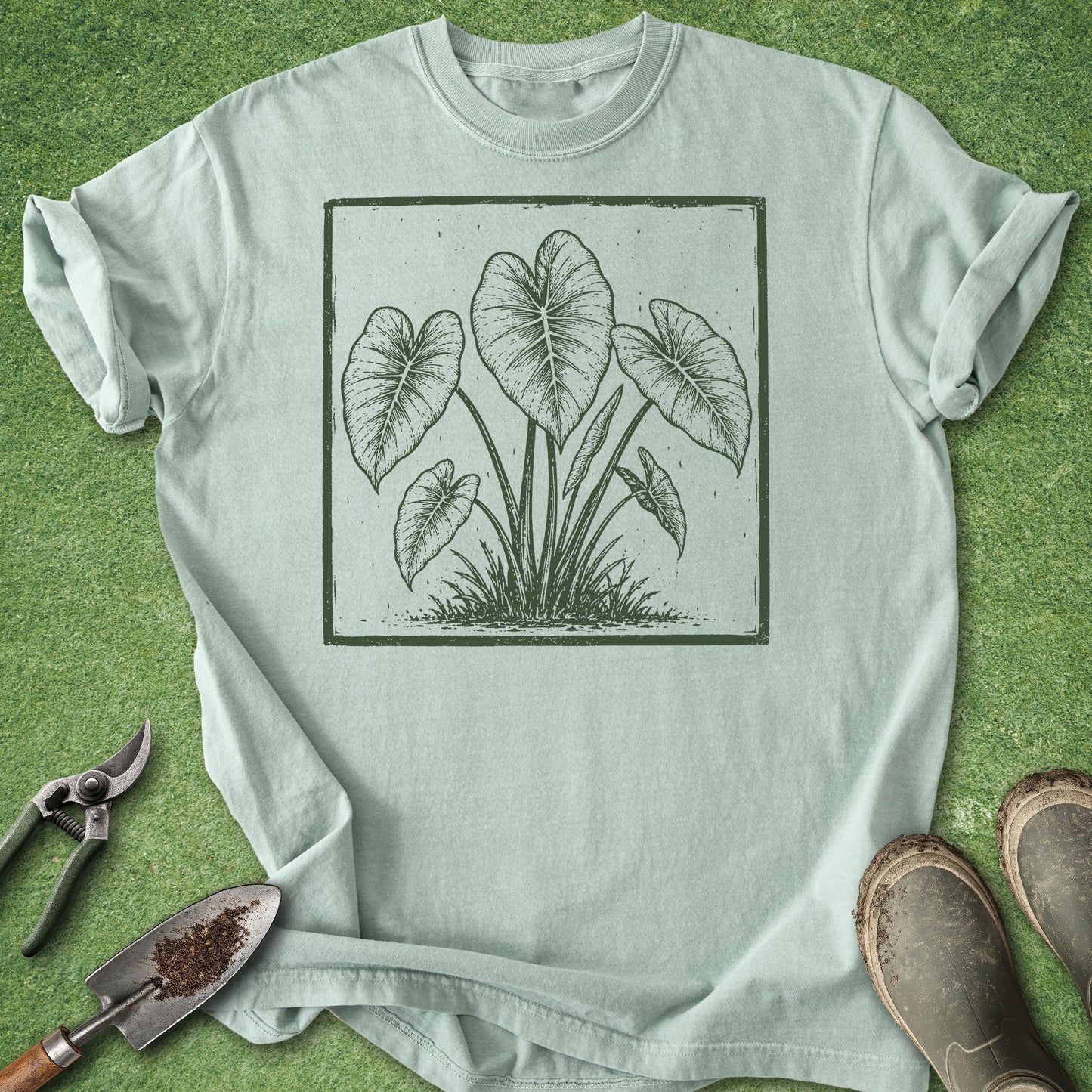 A light sage green t-shirt with a dark green botanical leaf graphic, laid flat on green grass with gardening tools (shears, trowel) and boots nearby.