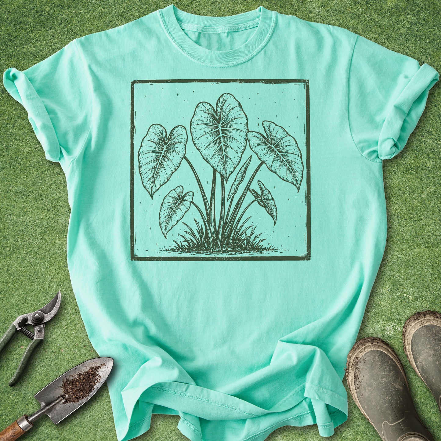A mint green short-sleeve t-shirt with a dark green botanical leaf graphic, laid flat on a green grass-like surface with gardening tools and boots nearby.
