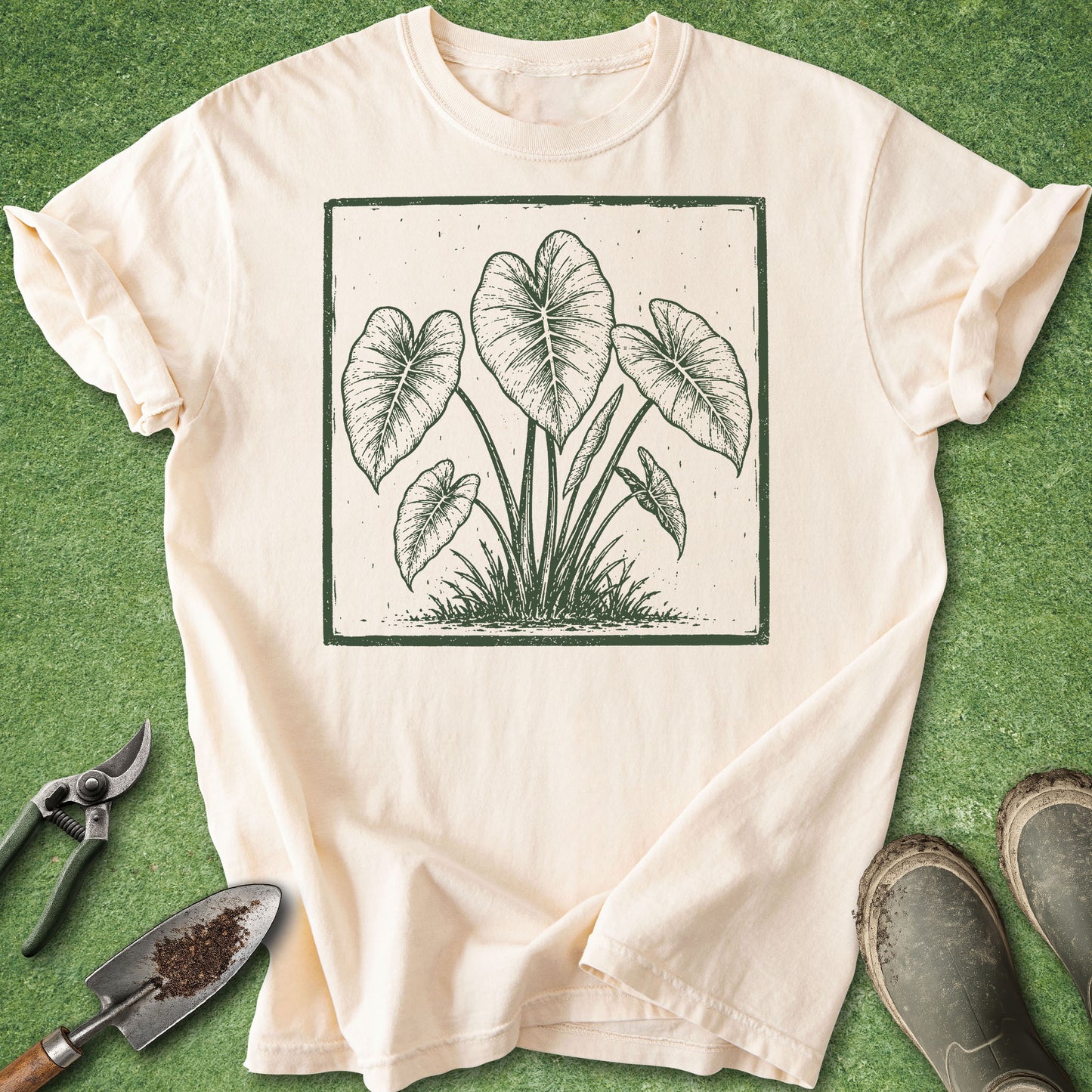 An off-white short-sleeve t-shirt with a dark green botanical leaf graphic, laid flat on a green grass-like surface with gardening props including pruning shears, a trowel, and boots.