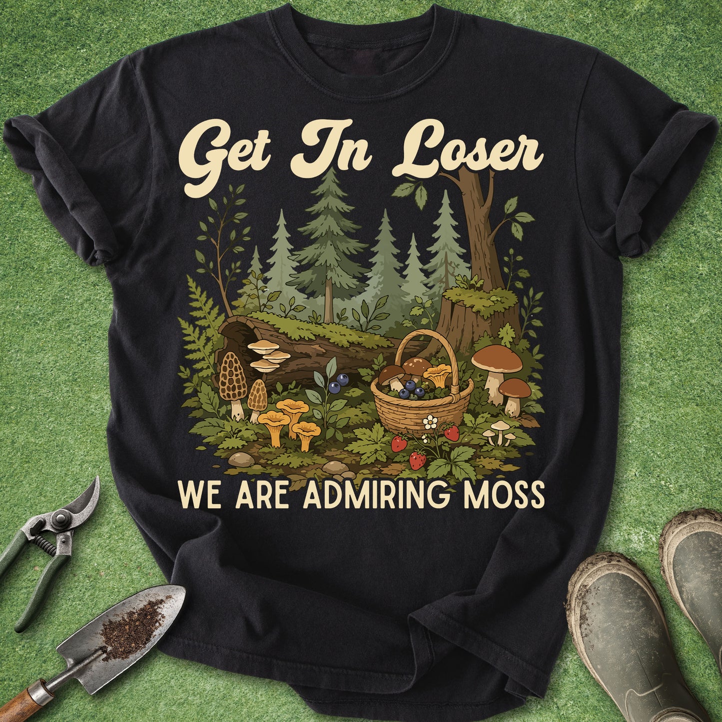 A black short-sleeve graphic T-shirt laid flat on a green grass-like surface, with a forest and mushroom illustration on the front and the text 'Get In Loser WE ARE ADMIRING MOSS.' Gardening tools and part of a pair of boots are placed around the shirt as styling props.