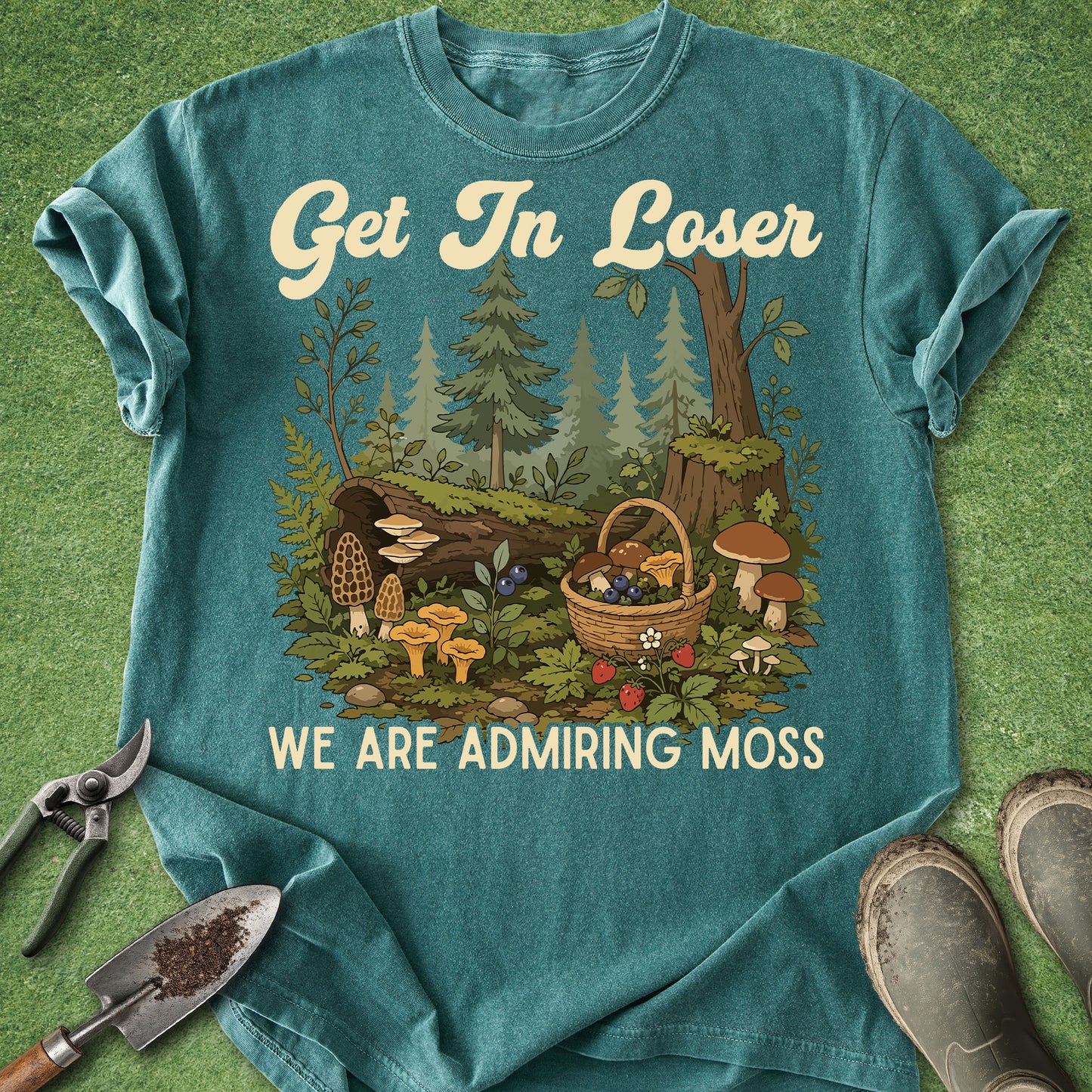 A teal graphic t-shirt laid flat on a green grass-like surface, featuring a forest illustration with mushrooms, a basket of berries, and the text 'Get In Loser WE ARE ADMIRING MOSS.' Gardening tools and boots are placed around the shirt as styling props.