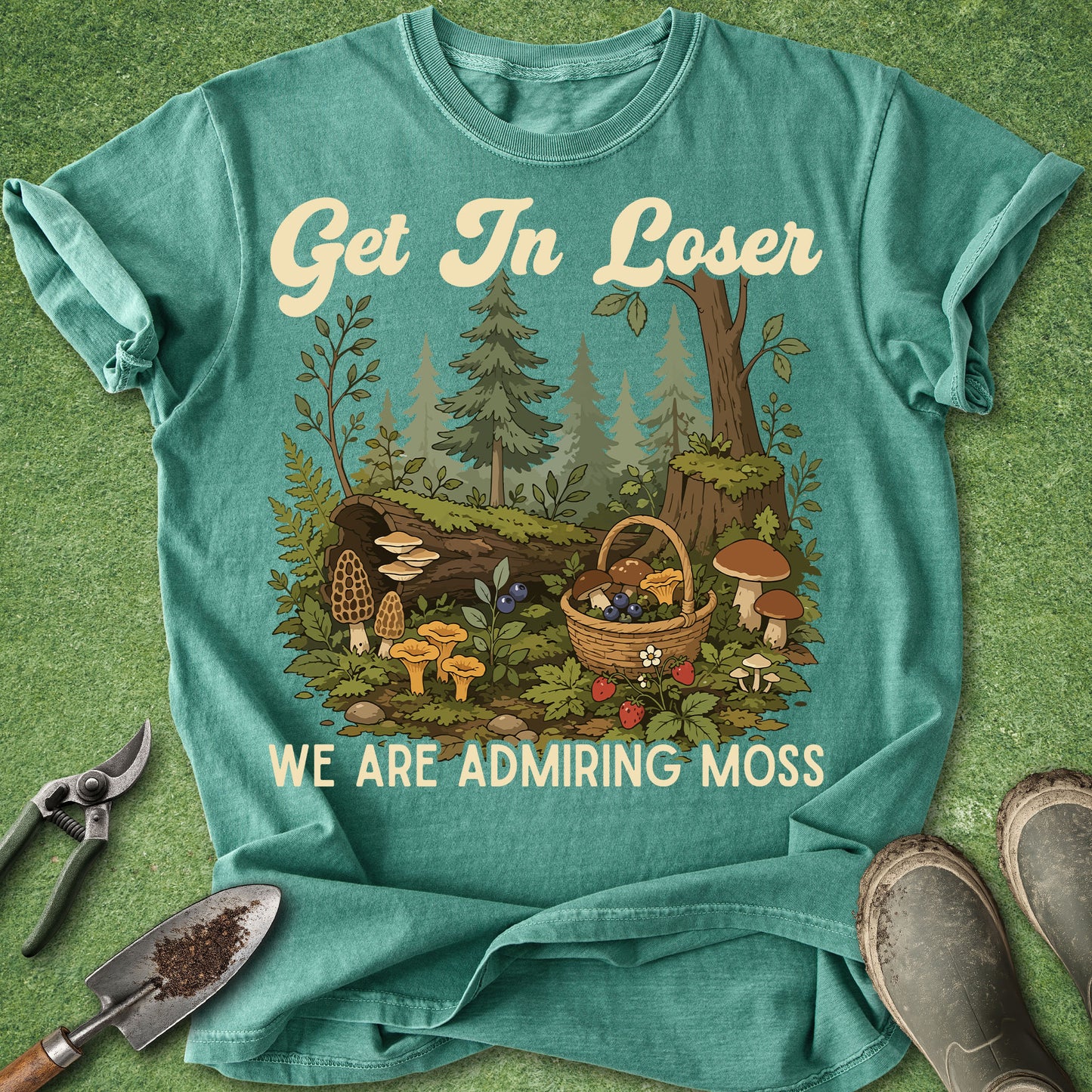 A teal graphic t-shirt laid flat on a green textured background, featuring a woodland forest scene with mushrooms, a basket, and the text 'Get In Loser WE ARE ADMIRING MOSS'. Gardening tools and boots are placed around the shirt as props.