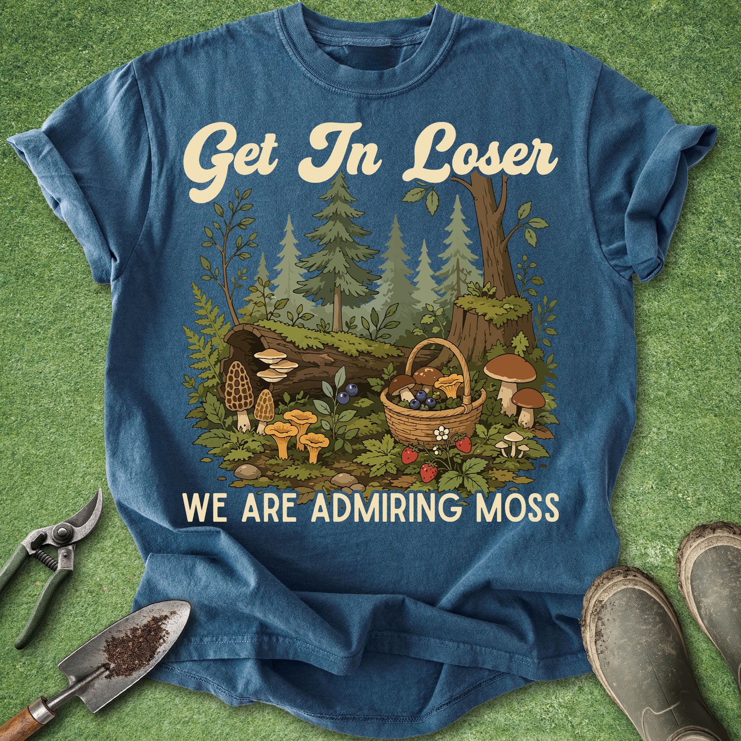 A blue graphic t-shirt laid flat on a green grass-like surface, with gardening tools and boots around it. The shirt has a forest scene printed on the front with mushrooms, a basket, and the text 'Get In Loser WE ARE ADMIRING MOSS'.