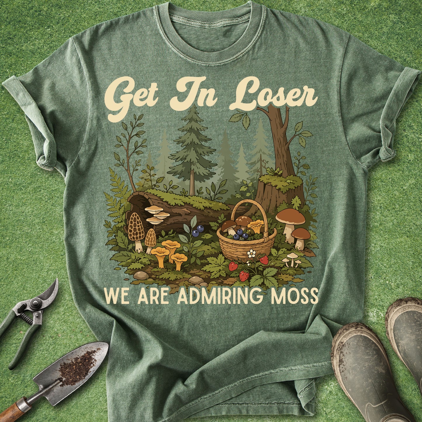 A green graphic t-shirt laid flat on a grassy background with gardening props like pruning shears, a trowel, and boots. The shirt features a woodland illustration of a mushroom basket and the text 'Get In Loser WE ARE ADMIRING MOSS'.