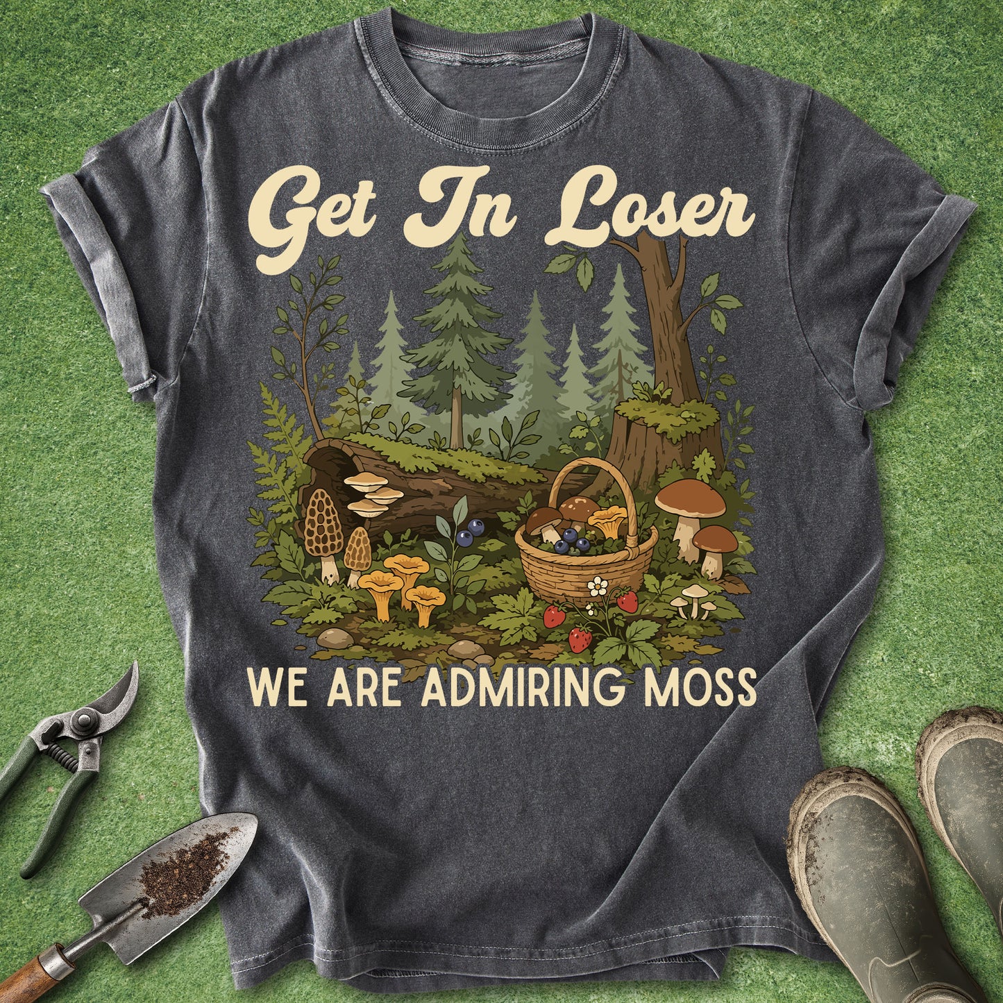 A dark charcoal gray graphic t-shirt laid flat on a green grass-like surface, surrounded by gardening props like pruning shears, a trowel, and boots. The shirt features a detailed forest illustration with mushrooms, a basket, and the text 'Get In Loser WE ARE ADMIRING MOSS'.
