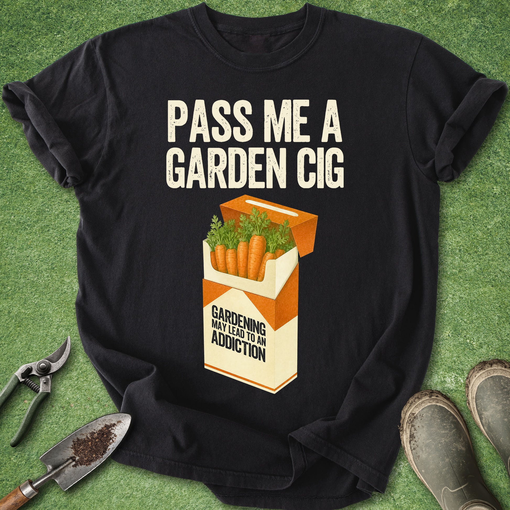 A black graphic t-shirt laid flat on green grass, featuring the text 'PASS ME A GARDEN CIG' above an illustrated orange and cream cigarette pack filled with carrots and green leaves. Gardening tools and boots are placed around the shirt as props.