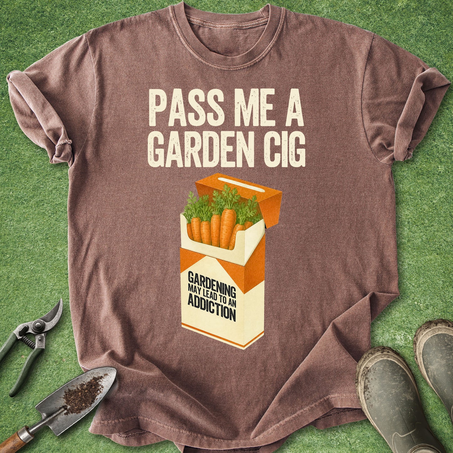 A brown short-sleeve graphic T-shirt laid flat on green grass-like background, with rolled sleeves and a printed design reading 'PASS ME A GARDEN CIG' above an orange-and-cream cigarette pack illustration filled with carrots. Gardening tools and boots are partially visible around the shirt.