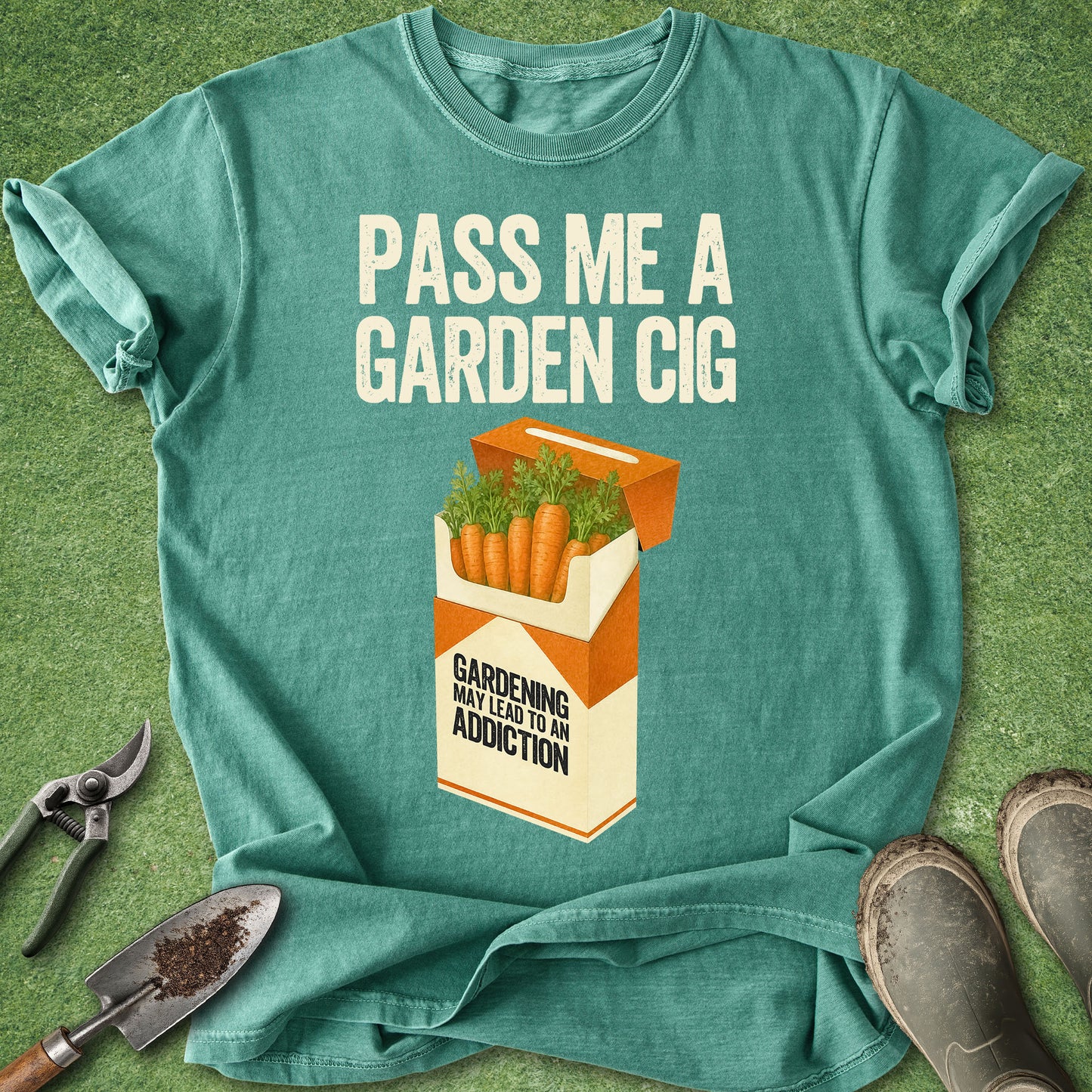 A teal graphic t-shirt laid flat on a green grass-like surface, with gardening tools and boots around it. The shirt has a humorous print that reads 'PASS ME A GARDEN CIG' above an illustrated orange-and-cream cigarette pack filled with carrots, with the text 'GARDENING MAY LEAD TO AN ADDICTION.