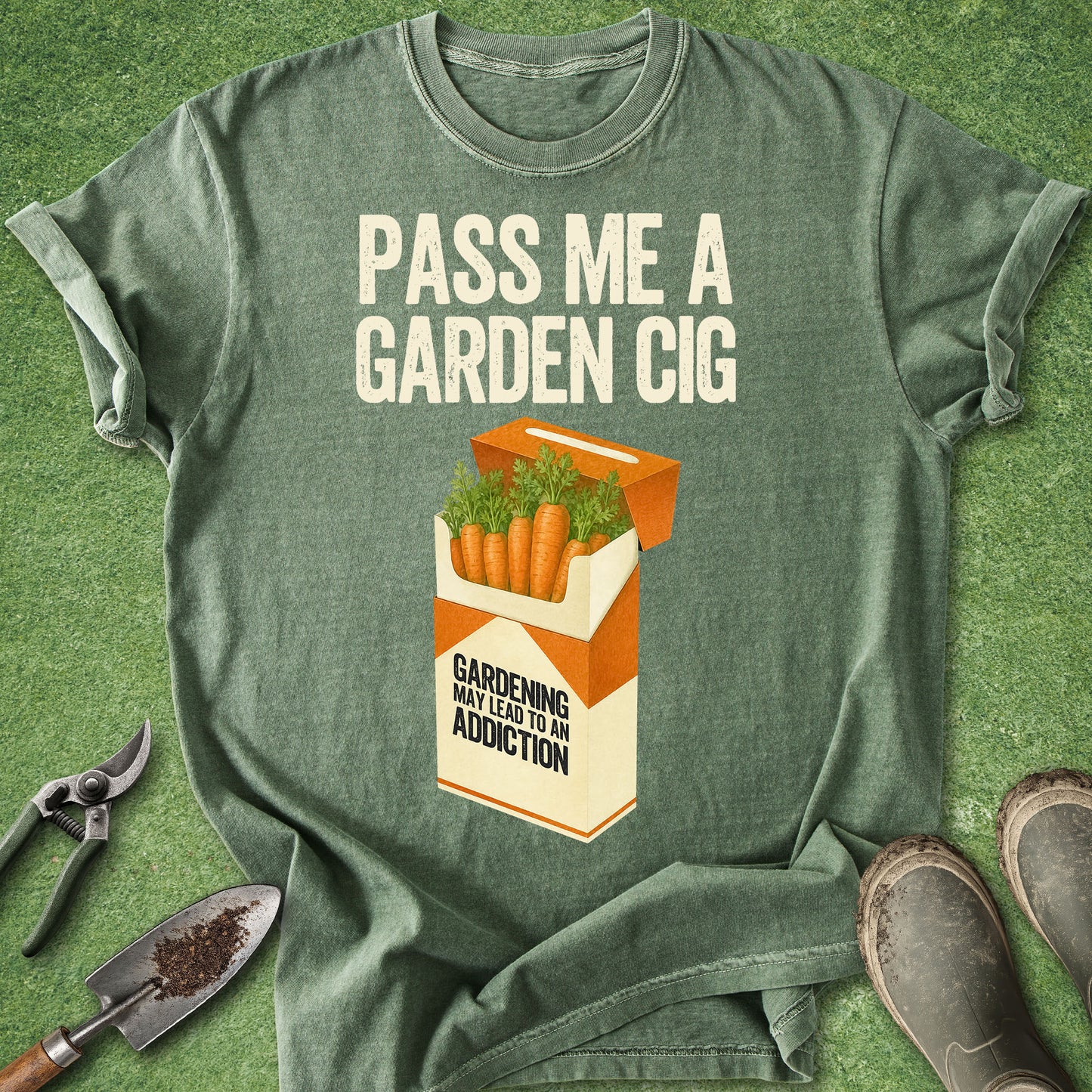Comfort Colors moss t-shirt featuring the phrase "Pass Me a Garden Cig" above a humorous carrot cigarette pack illustration with the warning "Gardening May Lead To An Addiction."