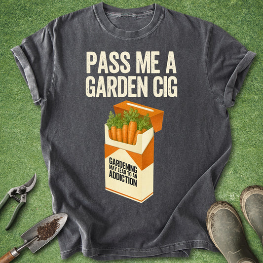 A dark gray graphic t-shirt laid flat on artificial green grass, styled with gardening tools and boots. The shirt features a humorous graphic of a cigarette pack filled with carrots and green leaves, with the text 'PASS ME A GARDEN CIG' and 'GARDENING MAY LEAD TO AN ADDICTION'.