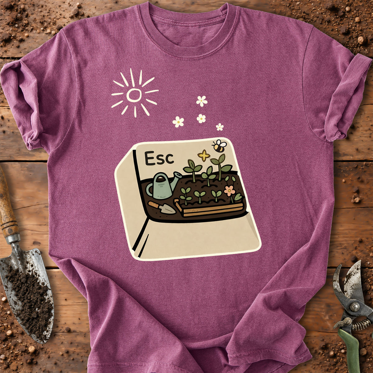 A heathered purple short-sleeve t-shirt laid flat on a rustic wooden surface with gardening tools. The shirt features a graphic print of a garden bed with a watering can, flowers, a bee, and a sun, with the letter 'Esc' printed on the top left of the graphic.