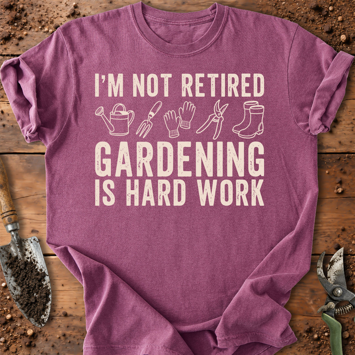A heathered purple short-sleeve t-shirt with a cream-colored graphic that reads 'I'M NOT RETIRED GARDENING IS HARD WORK' with simple gardening tool illustrations. The shirt is laid flat on a rustic wooden surface surrounded by gardening props like a trowel, soil, and pruning shears.