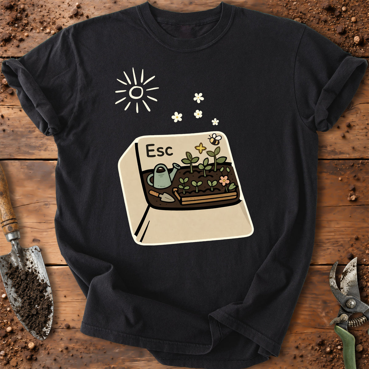 A black short-sleeve graphic T-shirt laid flat on a rustic wooden surface with gardening props like a trowel and pruning shears. The shirt features a large front print of a beige computer keyboard key with the word 'Esc' and a gardening illustration including a sun, flowers, a watering can, and a planter with small plants.