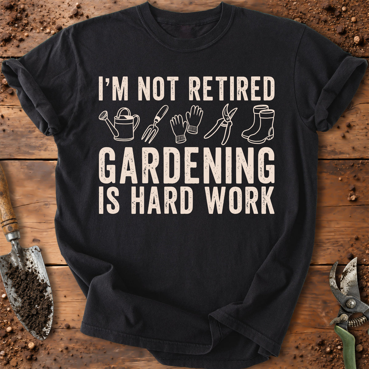 A black short-sleeve graphic T-shirt laid flat on a rustic wooden surface with gardening tools around it. The shirt has a distressed cream print that reads “I’M NOT RETIRED GARDENING IS HARD WORK” with small icons of a watering can, rake, gloves, shears, and boots.
