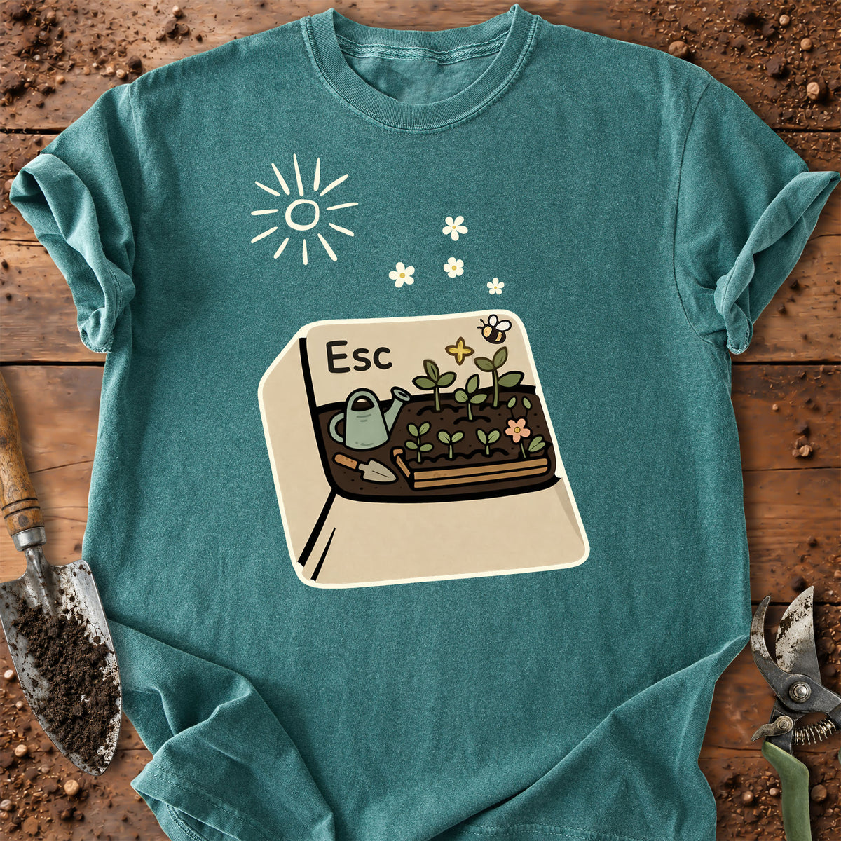 A teal short-sleeve t-shirt with a gardening-themed graphic print featuring a sun, flowers, and a planter box labeled 'Esc' with a watering can and bee. The shirt is laid flat on a rustic wooden surface with gardening tools like a trowel and pruning shears visible around the edges.