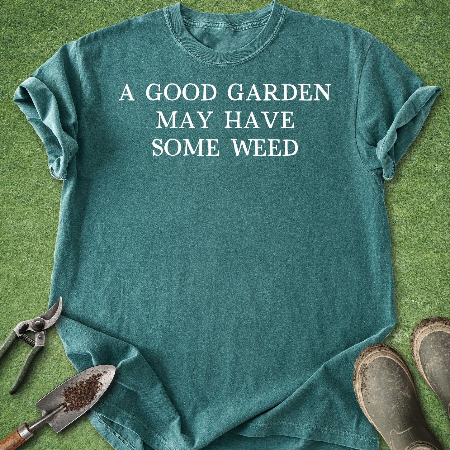 Teal t-shirt with 'A good garden may have some weed' text on grass with gardening tools.