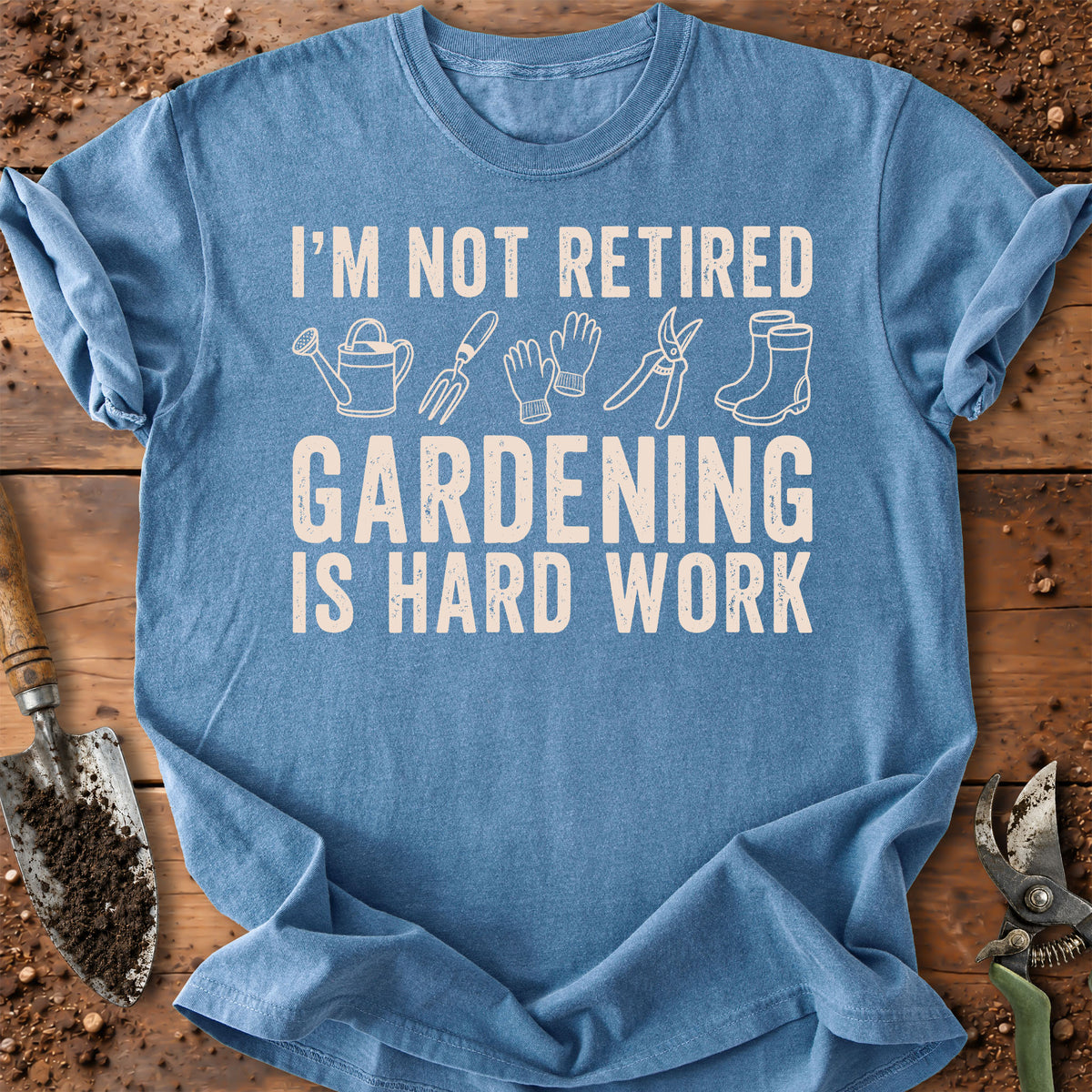 A light blue crewneck t-shirt with a white graphic text reading 'I'M NOT RETIRED GARDENING IS HARD WORK' accompanied by simple gardening tool illustrations. The shirt is laid flat on a rustic wooden surface surrounded by gardening props like a trowel and pruning shears.
