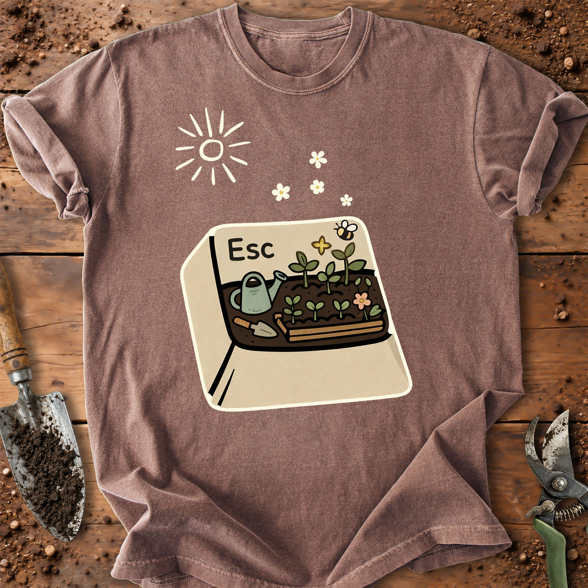 A mauve-brown short-sleeve t-shirt laid flat on a rustic wooden surface with gardening props. The shirt features a graphic of a computer keyboard key labeled 'Esc' containing a cartoon garden planter with a watering can, flowers, and a bee.