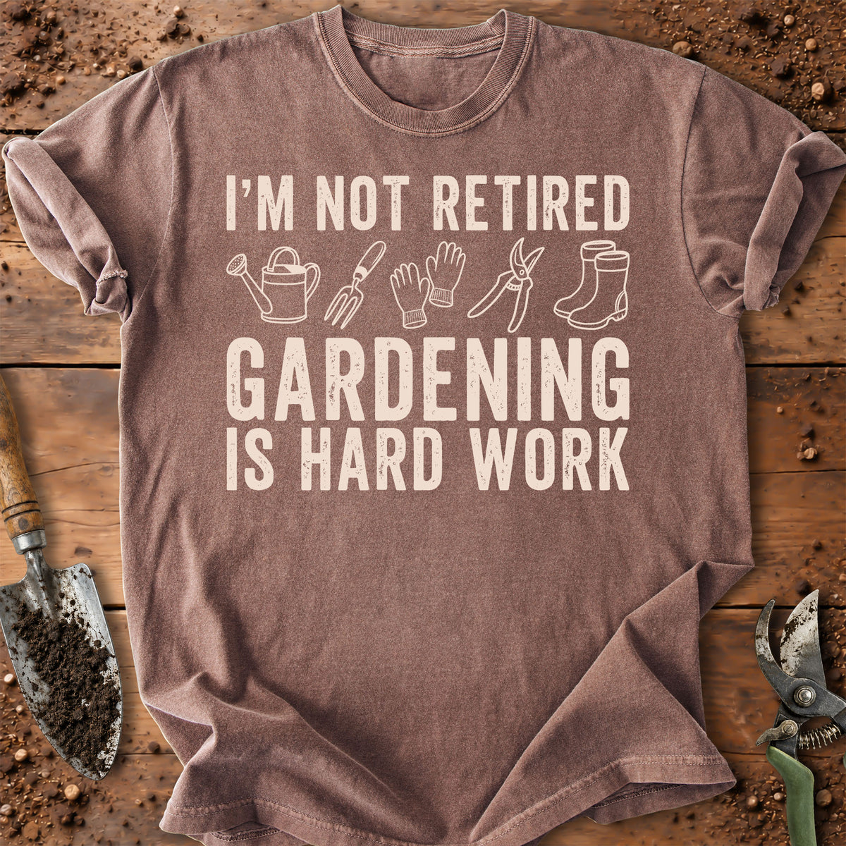 A heathered brown short-sleeve t-shirt with a white graphic print that reads 'I'M NOT RETIRED GARDENING IS HARD WORK' with simple gardening tool icons. The shirt is laid flat on a rustic wooden surface surrounded by gardening props like a trowel, soil, and pruning shears.