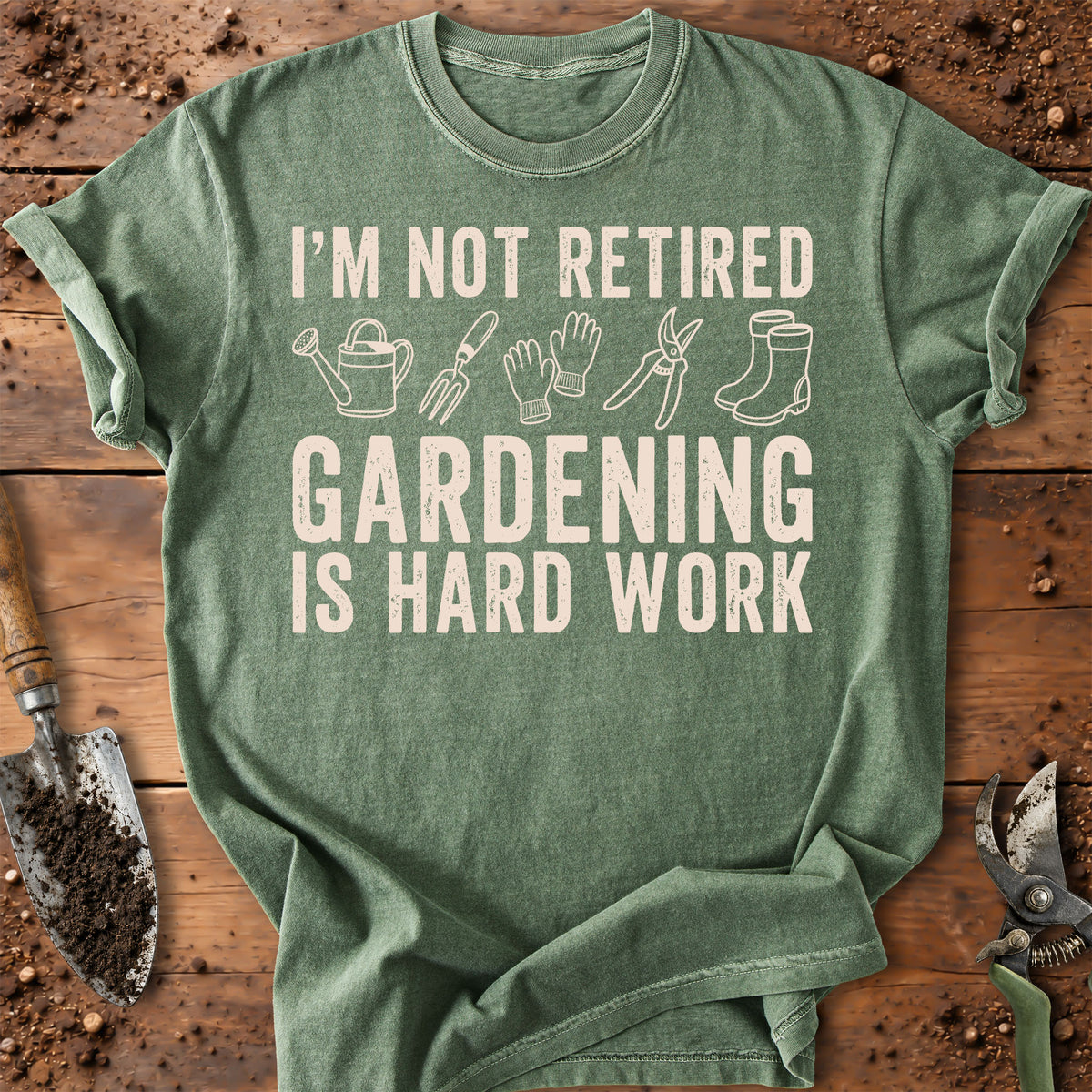 A heather sage green t-shirt with a cream-colored graphic that reads 'I'M NOT RETIRED GARDENING IS HARD WORK' accompanied by simple line drawings of gardening tools (watering can, rake, gloves, pruning shears, boots). The t-shirt is laid flat on a rustic wooden background with gardening props like a trowel and pruning shears visible around the edges.
