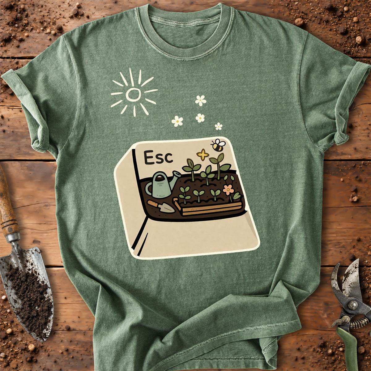 A sage green graphic t-shirt laid flat on a rustic wooden background with gardening props like a trowel and pruning shears. The graphic features a beige card with a gardening illustration labeled 'Esc' and a sun icon above it.