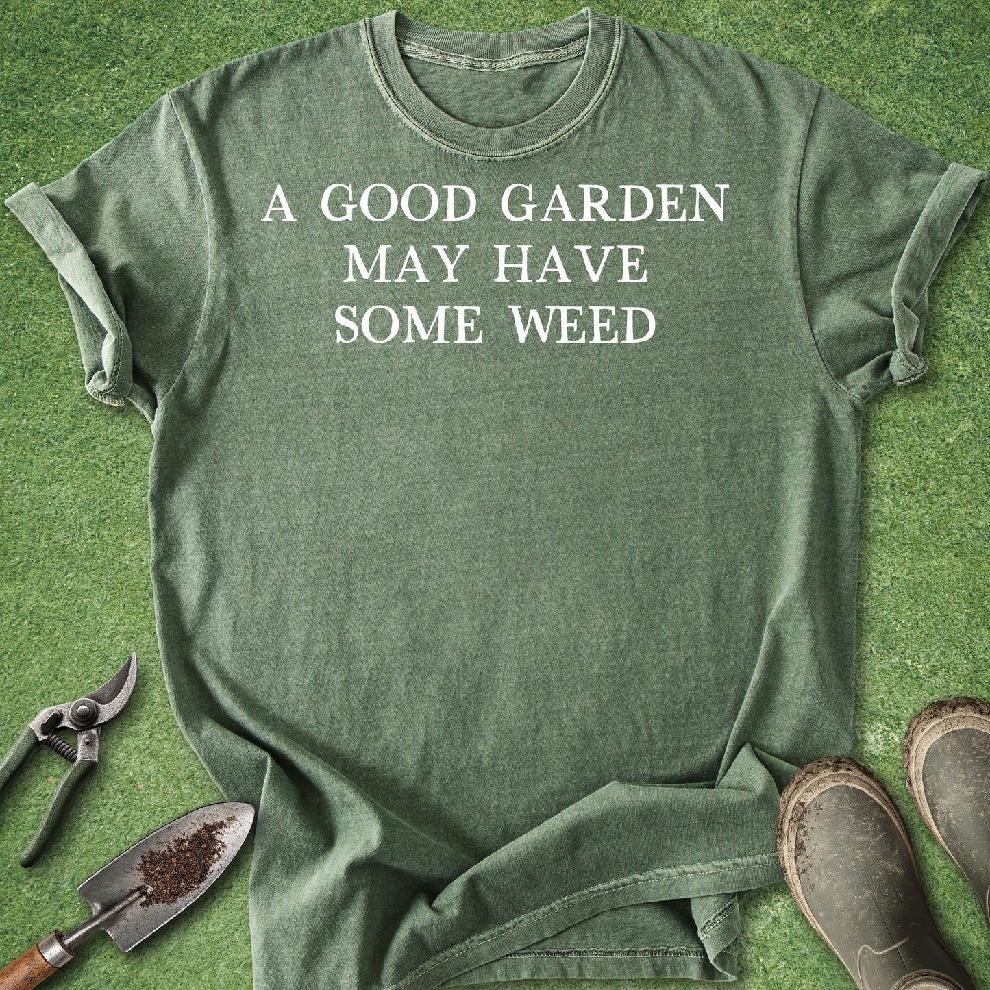 Green t-shirt with 'A Good Garden May Have Some Weed' text on a grass background with gardening tools.