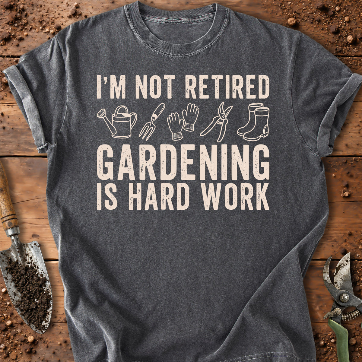 A dark gray graphic t-shirt laid flat on a rustic wooden surface with gardening tools around it. The shirt features a cream-colored text graphic that reads 'I'M NOT RETIRED GARDENING IS HARD WORK' with simple line drawings of a watering can, rake, gloves, pruning shears, and boots.