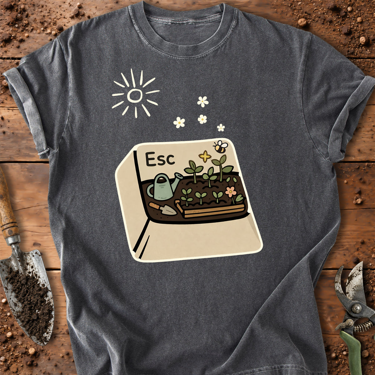 A dark gray graphic t-shirt laid flat on a rustic wooden surface with gardening props like a trowel and pruning shears. The shirt features a central graphic of a garden bed with a watering can, flowers, a bee, and the word 'Esc' inside a beige key illustration, alongside a simple white sun graphic.