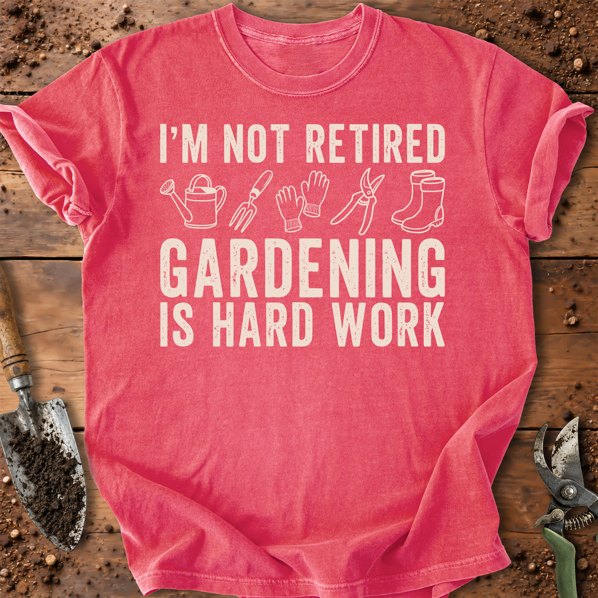 A coral pink short-sleeve t-shirt with a white graphic text reading 'I'M NOT RETIRED GARDENING IS HARD WORK' accompanied by simple gardening tool illustrations. The shirt is laid flat on a rustic wooden surface with gardening props like a trowel and pruning shears visible around it.