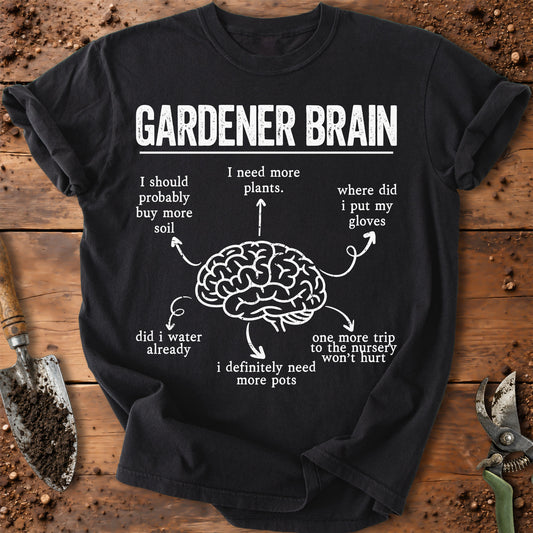 A black short-sleeve graphic T-shirt laid flat on a rustic wooden surface with gardening props. The shirt has a white printed design reading 'GARDENER BRAIN' above a brain illustration with humorous arrows and text pointing to it.