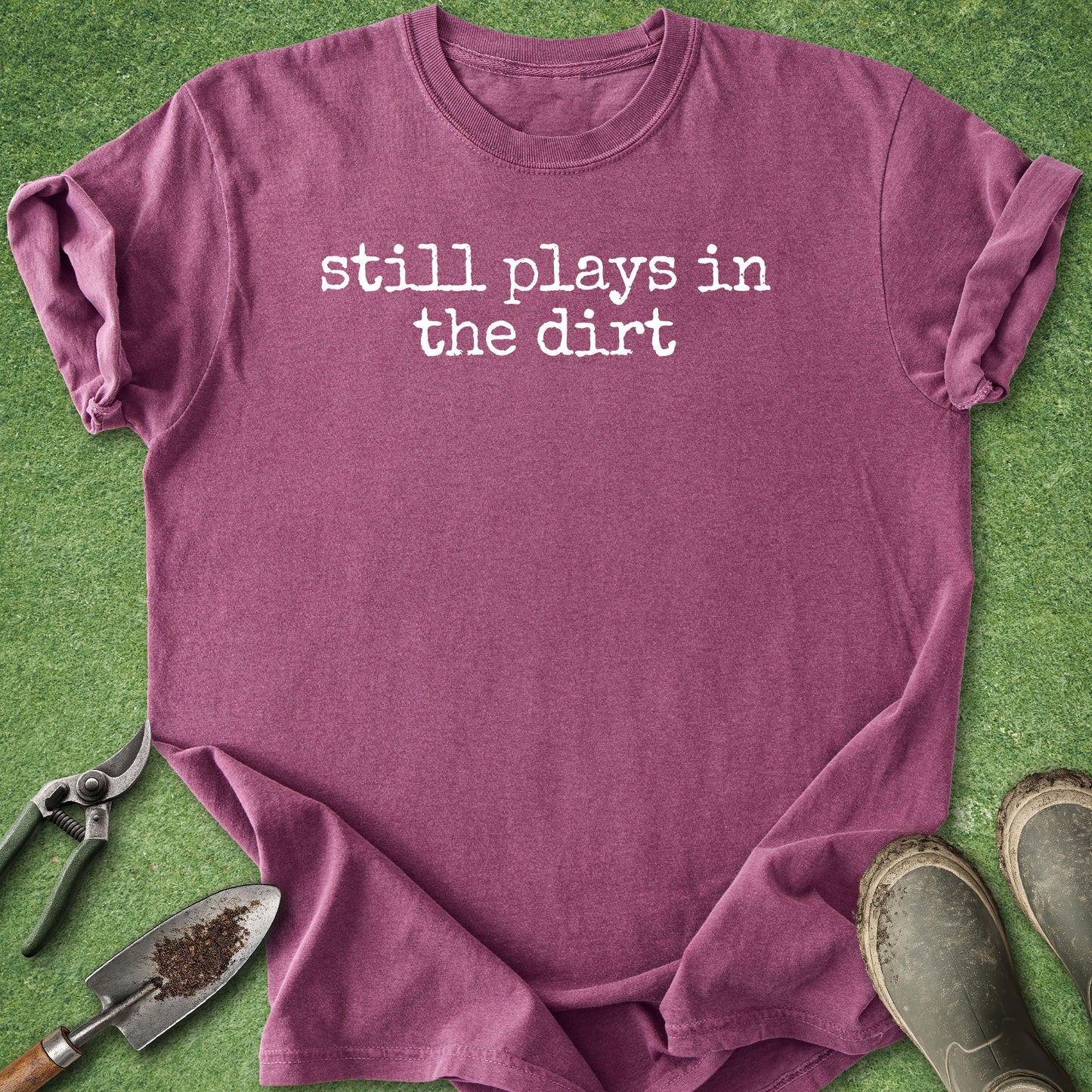 A heathered mauve-purple t-shirt with the phrase 'still plays in the dirt' printed in white typewriter-style text across the chest. The shirt is laid flat on green grass with gardening props, including a trowel, pruning shears, and muddy boots, visible around it.