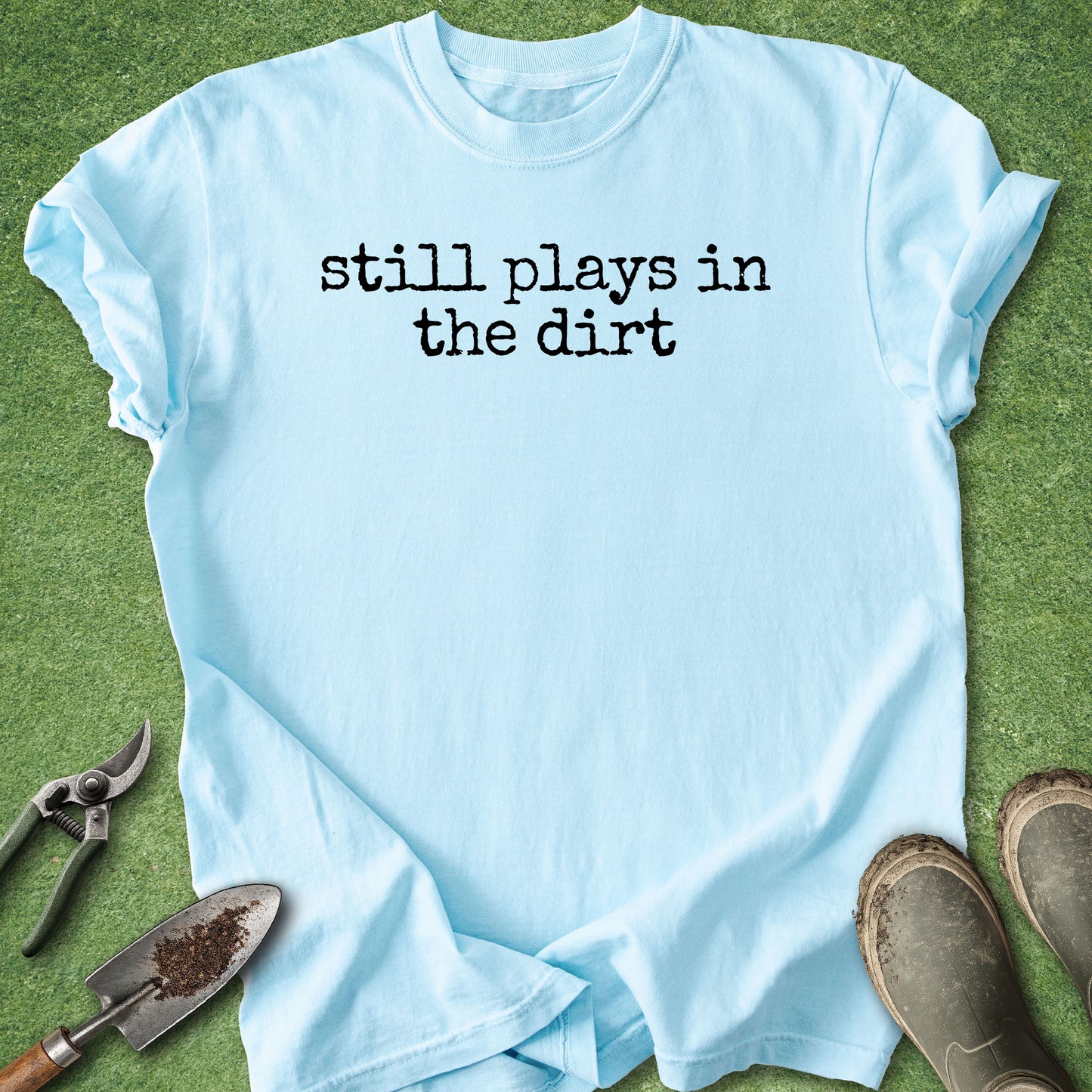 A light blue crewneck t-shirt with the text 'still plays in the dirt' printed in black across the chest, laid flat on green grass with gardening tools and boots nearby.