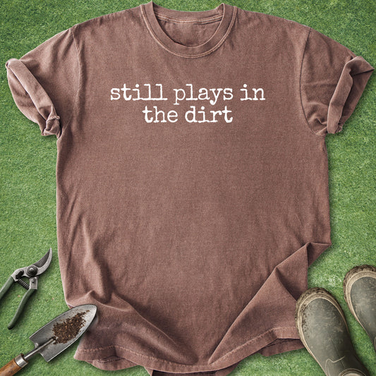 A brown short-sleeve t-shirt with the white text 'still plays in the dirt' printed on the chest, laid flat on green grass with gardening tools and boots around it.