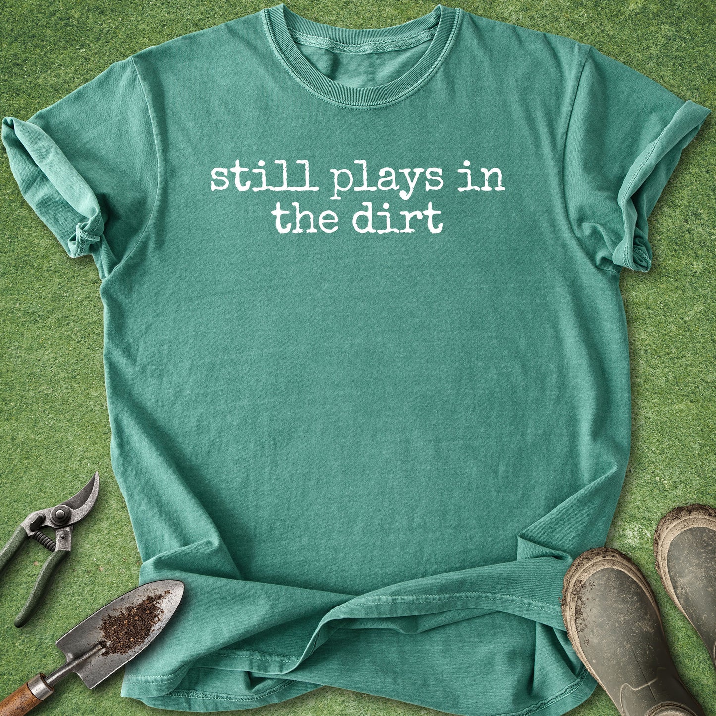 A teal green short-sleeve t-shirt with the phrase 'still plays in the dirt' printed in white lowercase letters across the chest. The shirt is laid flat on green grass, styled with gardening tools (a trowel, pruning shears, and boots) around it.