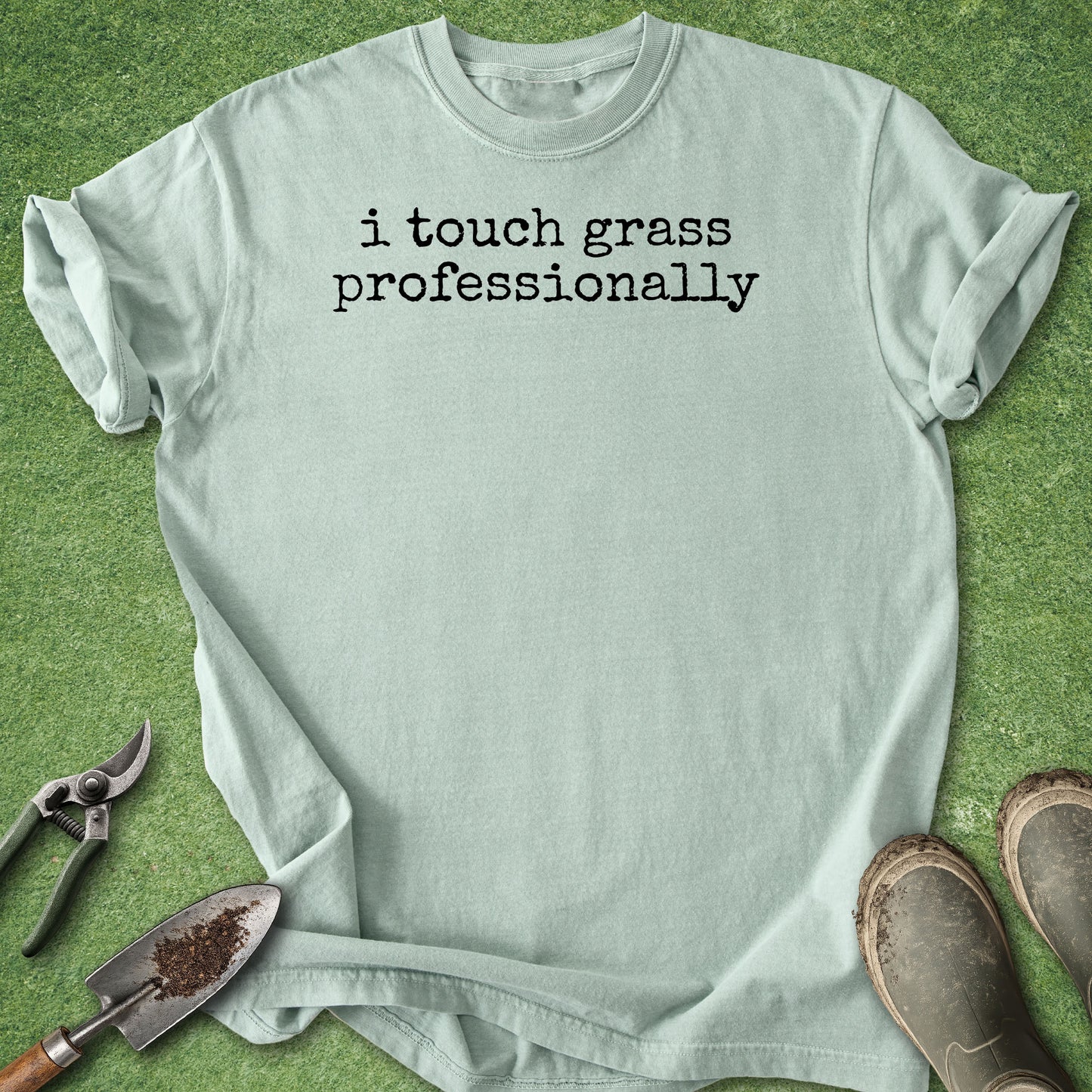 A light sage green t-shirt with the text 'i touch grass professionally' printed in black typewriter font, laid flat on green grass with gardening tools and boots nearby.