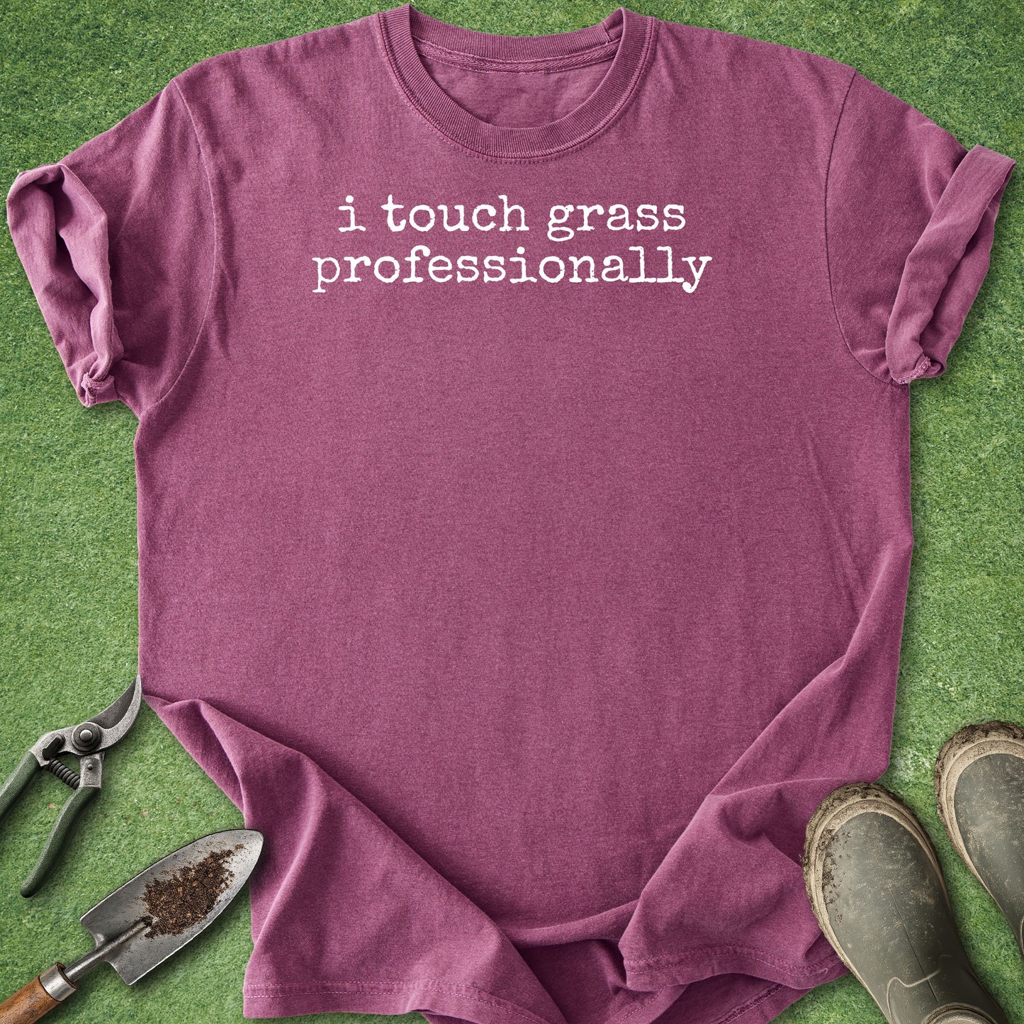 A heathered purple short-sleeve t-shirt with the phrase 'i touch grass professionally' printed in white lowercase letters across the chest. The shirt is laid flat on green artificial grass with gardening props, including pruning shears, a trowel, and boots, visible around it.
