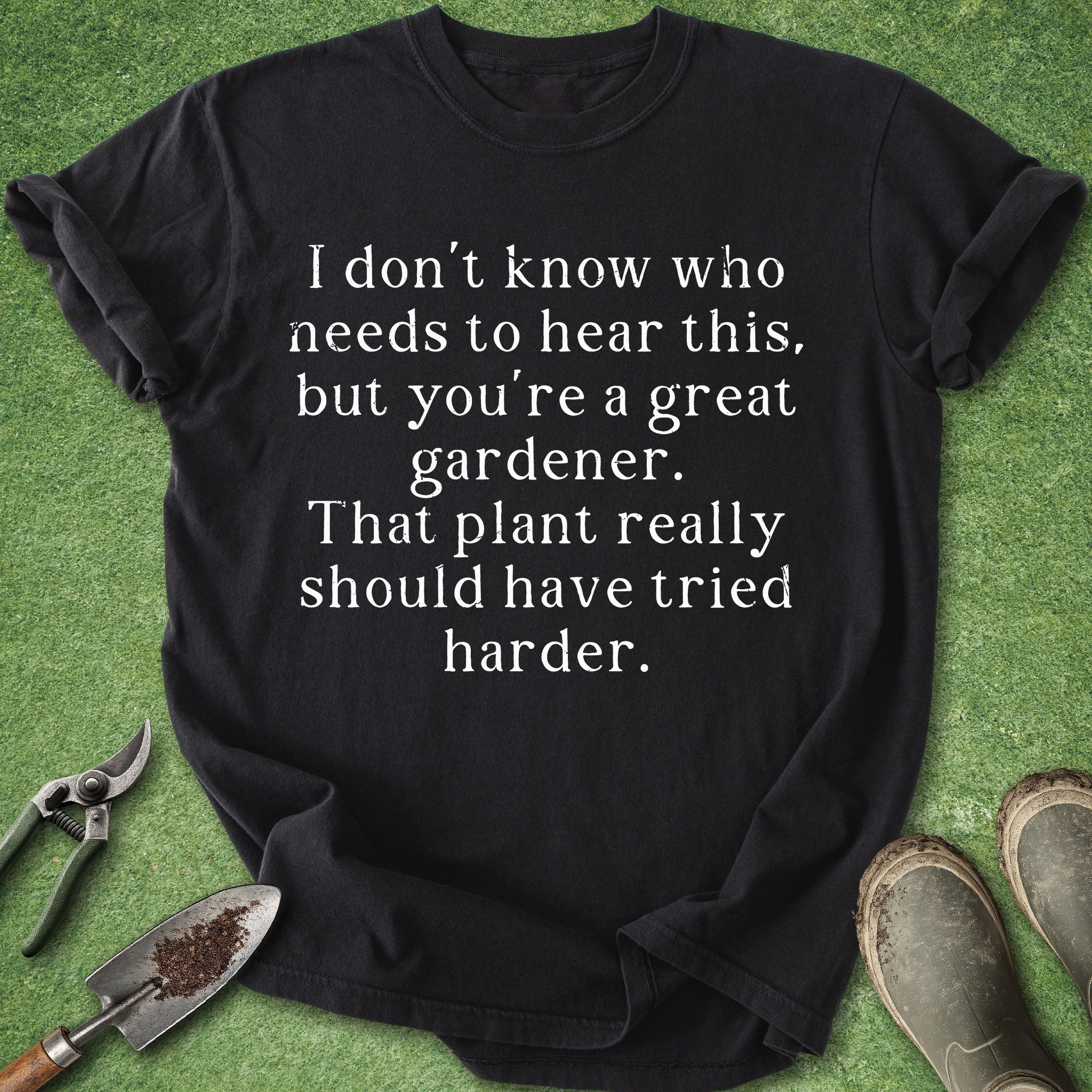 Black t-shirt with humorous gardening text on a green background with gardening tools.