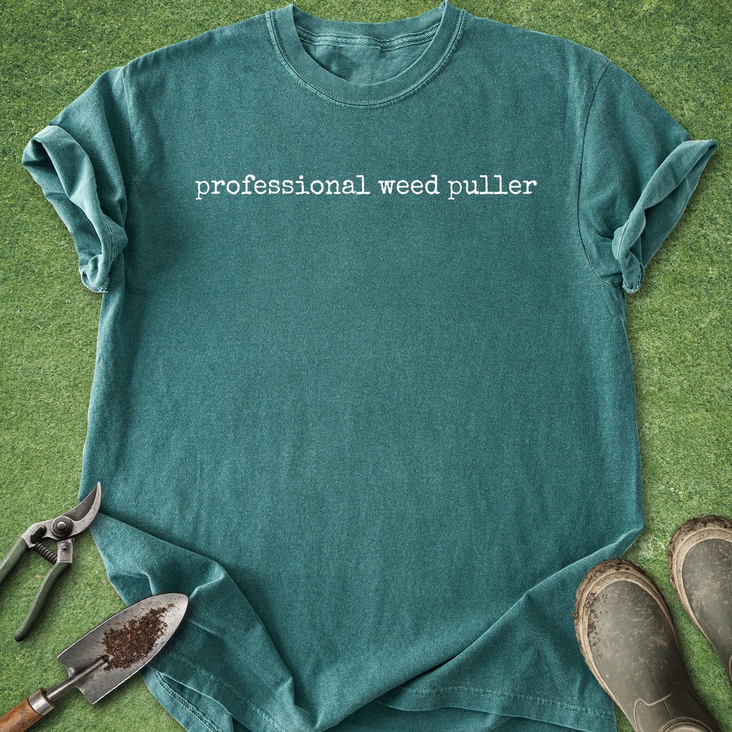 Teal t-shirt with 'professional weed puller' text on a grass background with gardening tools.