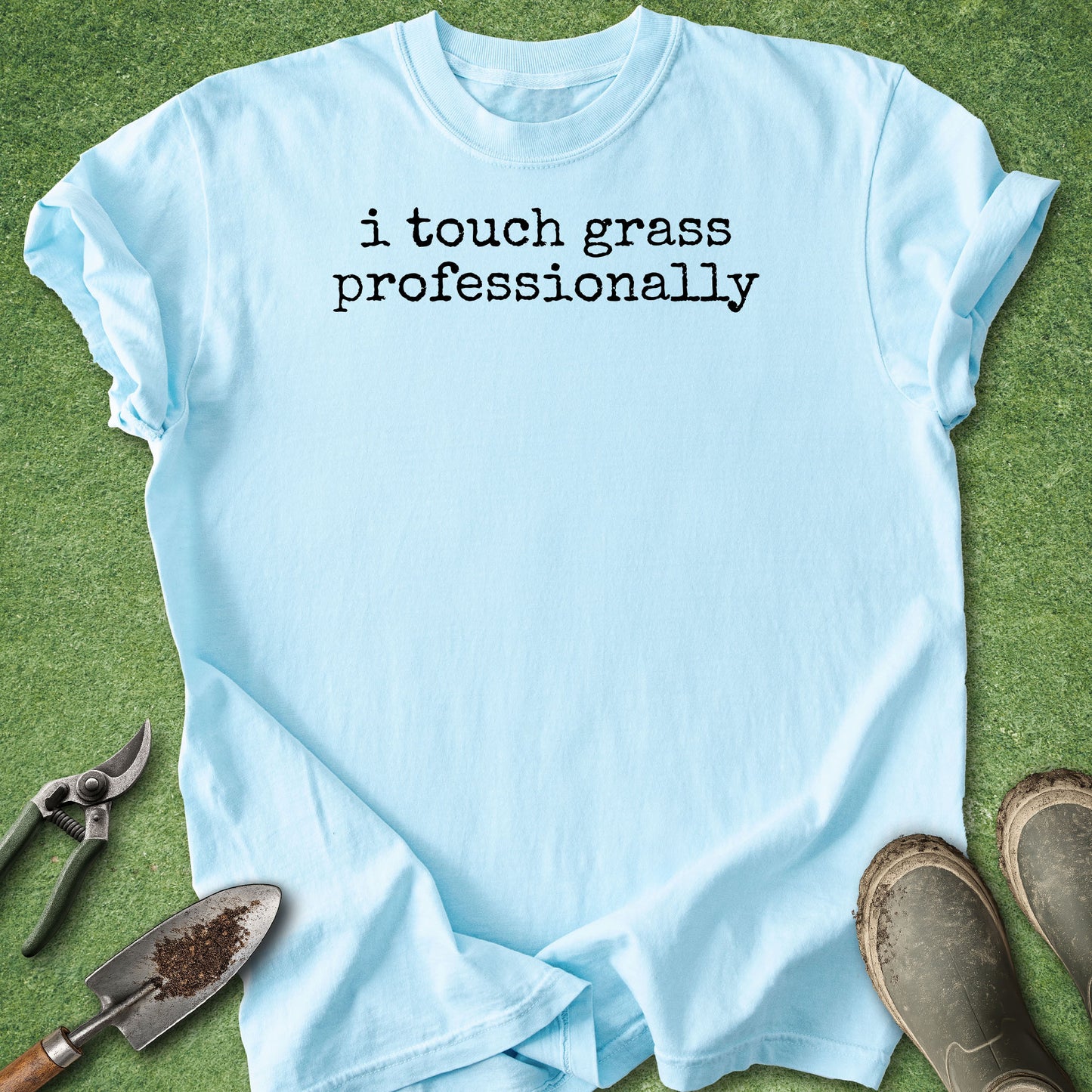 A light blue crewneck t-shirt with the text 'i touch grass professionally' printed in black on the chest, laid flat on green grass with gardening tools and boots around it.