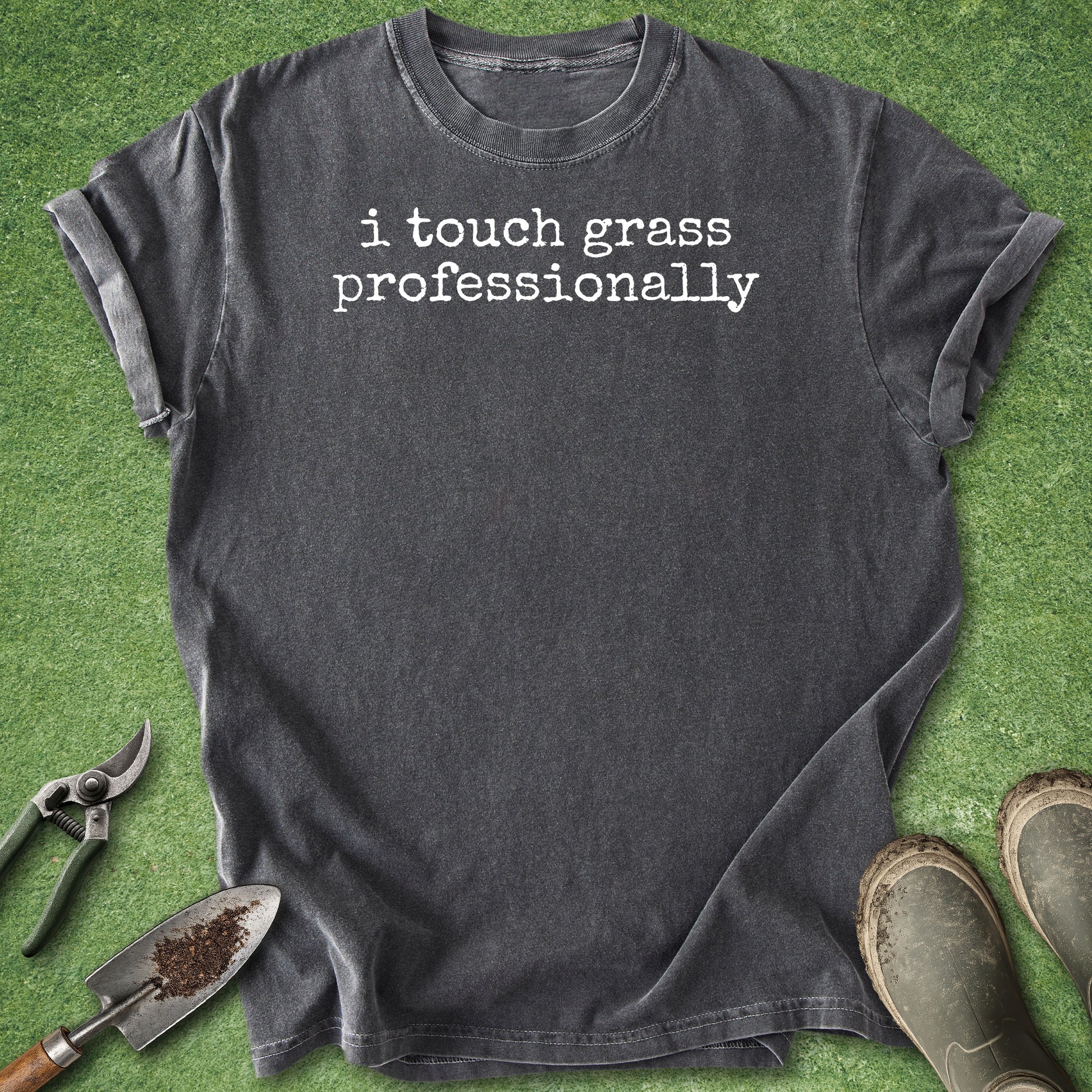 A dark charcoal gray graphic t-shirt with the text 'i touch grass professionally' printed in white typewriter font. The t-shirt is laid flat on green grass with gardening props, including pruning shears, a trowel with soil, and work boots, around it.