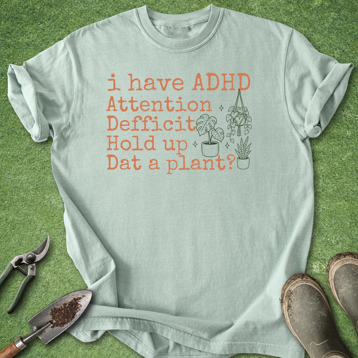 A light sage green t-shirt with a humorous graphic text reading 'i have ADHD Attention Deficit Hold up Dat a plant?' in orange, styled in a flat-lay on green grass with gardening tools and boots.