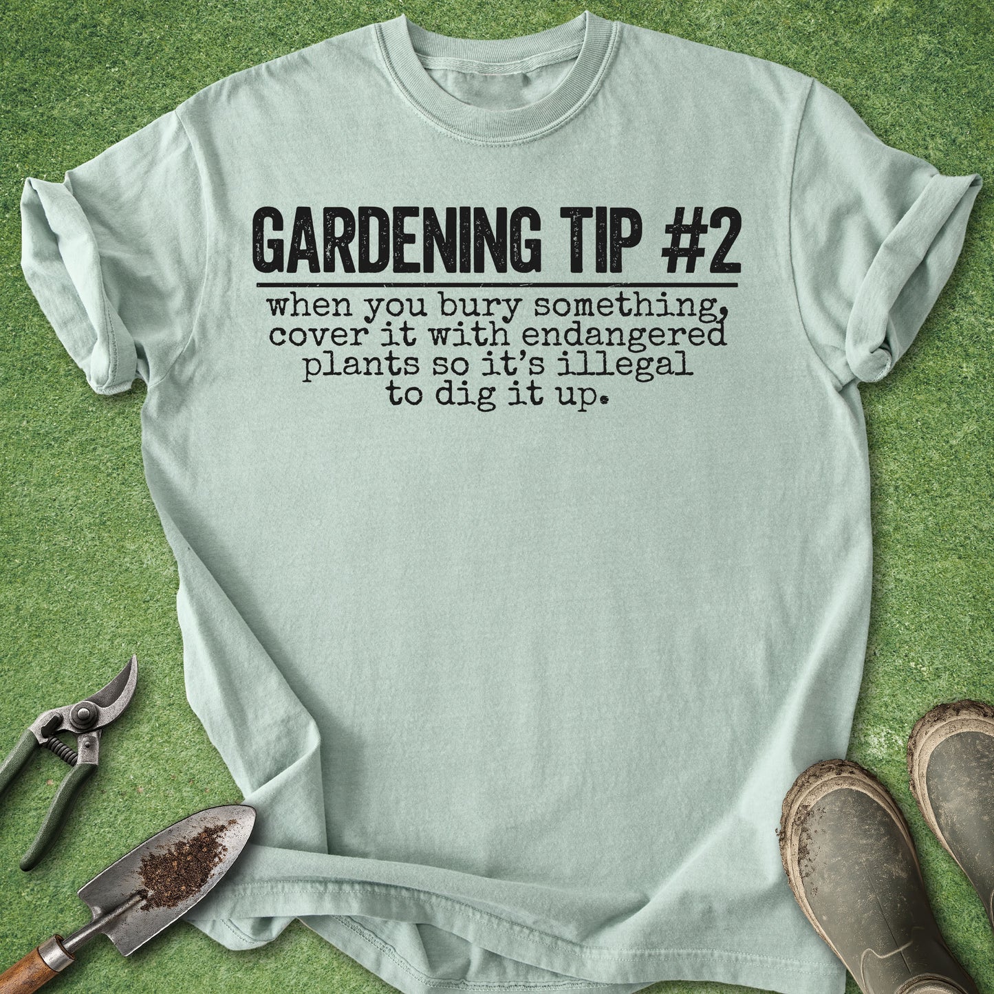 A light sage green graphic t-shirt laid flat on a green grass-like surface, with gardening tools (shears, trowel) and muddy boots around it. The shirt features black text reading 'GARDENING TIP #2 when you bury something, cover it with endangered plants so it's illegal to dig it up.