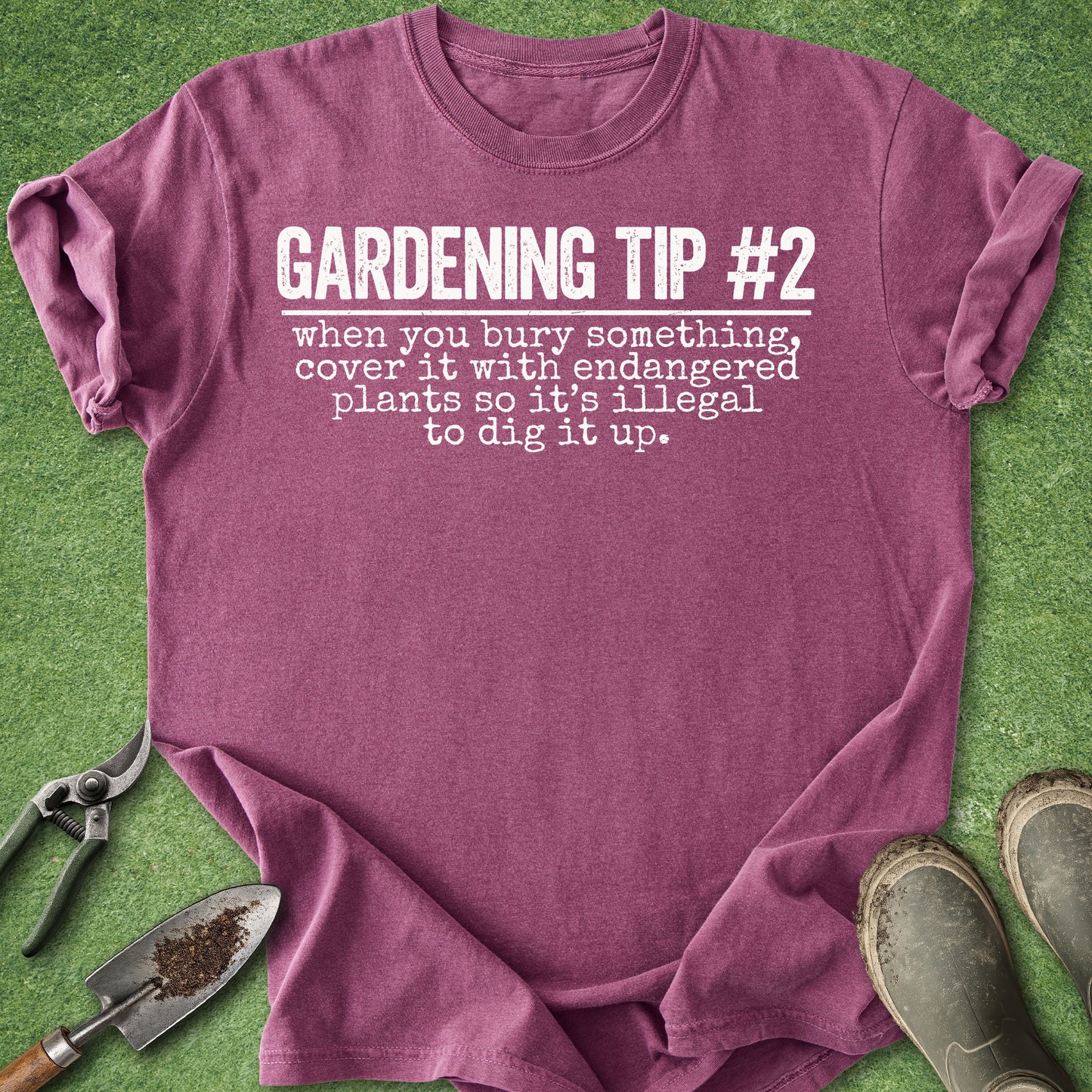A heathered mauve-purple t-shirt with white printed text reading 'GARDENING TIP #2 when you bury something, cover it with endangered plants so it's illegal to dig it up.' It is laid flat on green grass-like material with gardening props including pruning shears, a small trowel, and boots around it.
