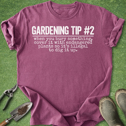 A heathered mauve-purple t-shirt with white printed text reading 'GARDENING TIP #2 when you bury something, cover it with endangered plants so it's illegal to dig it up.' It is laid flat on green grass-like material with gardening props including pruning shears, a small trowel, and boots around it.