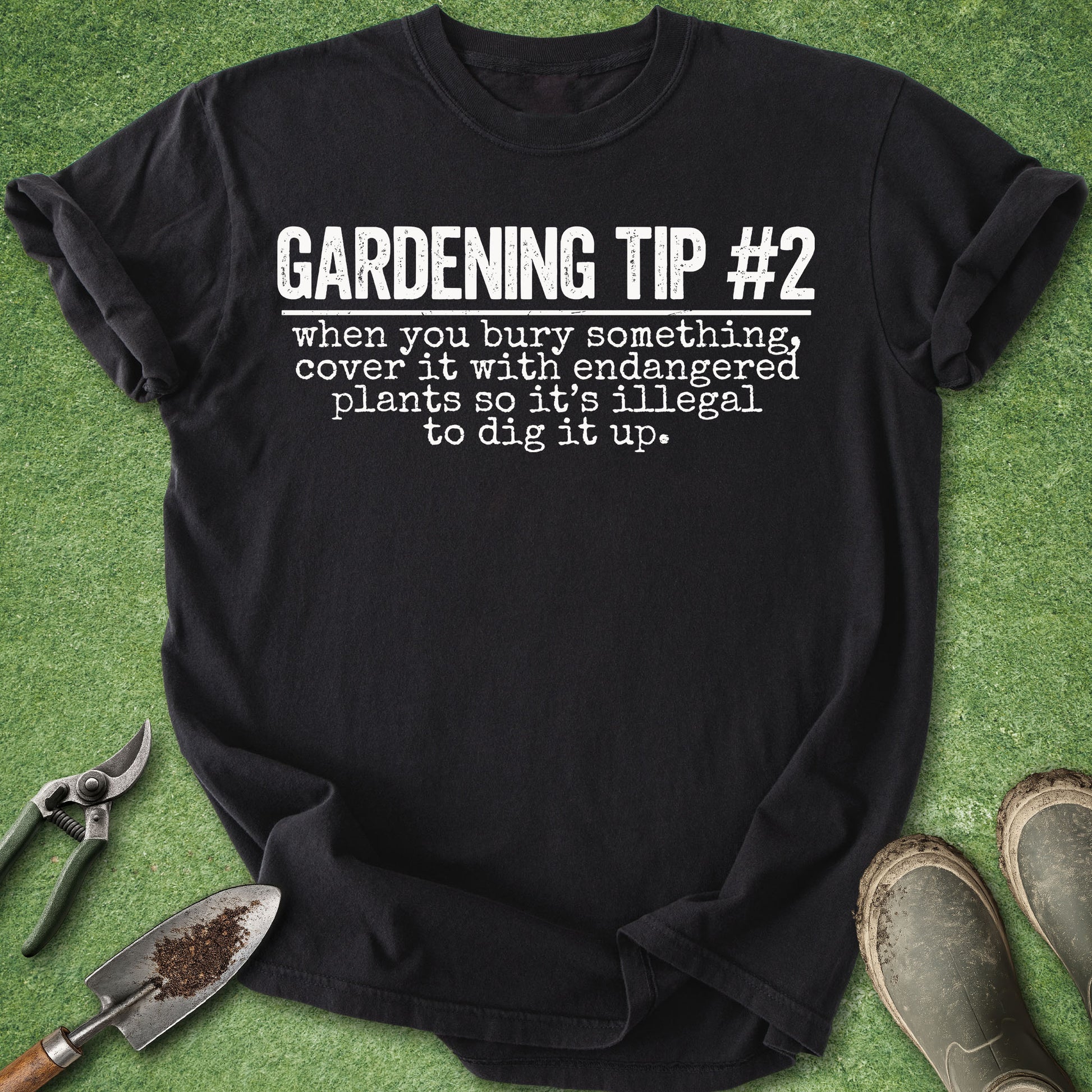 A black short-sleeve graphic T-shirt laid flat on green artificial grass, with gardening tools and muddy boots around it. The shirt reads “GARDENING TIP #2 when you bury something, cover it with endangered plants so it’s illegal to dig it up.