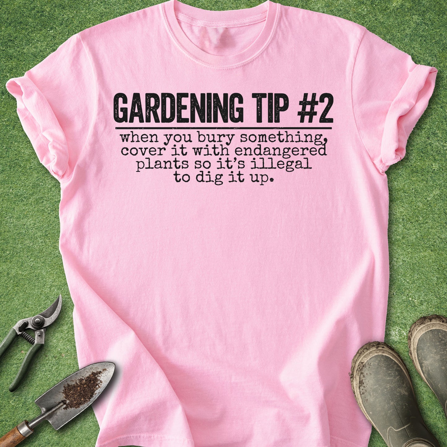 A light pink short-sleeve t-shirt with black printed text reading 'GARDENING TIP #2 when you bury something, cover it with endangered plants so it's illegal to dig it up.' The shirt is laid flat on green grass-like material with gardening props including pruning shears, a small trowel, and muddy boots around it.