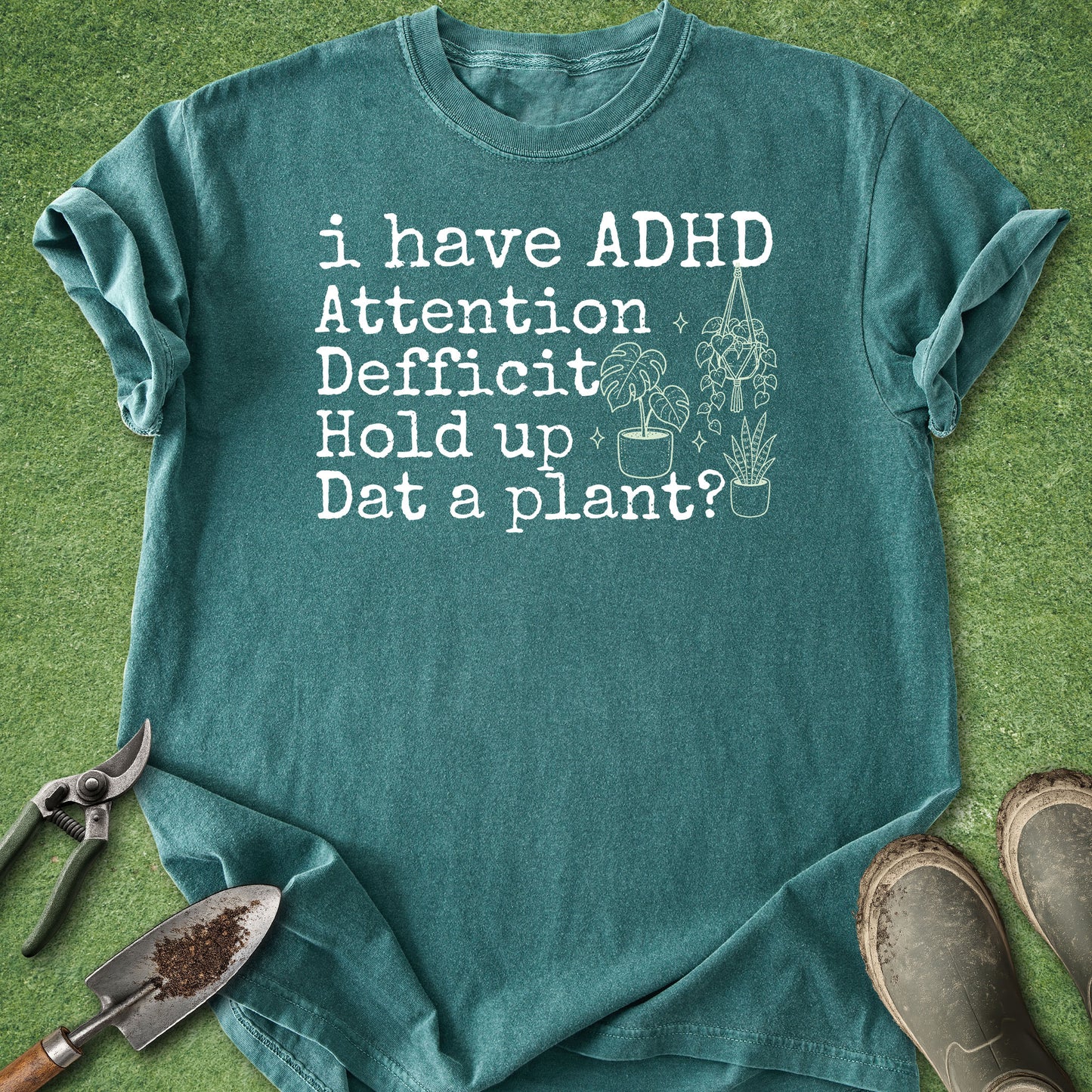 A teal graphic t-shirt laid flat on green grass with gardening props, including pruning shears, a trowel, and muddy boots. The shirt features white text that reads 'i have ADHD Attention Deficit Hold up Dat a plant?' alongside simple illustrations of potted plants.