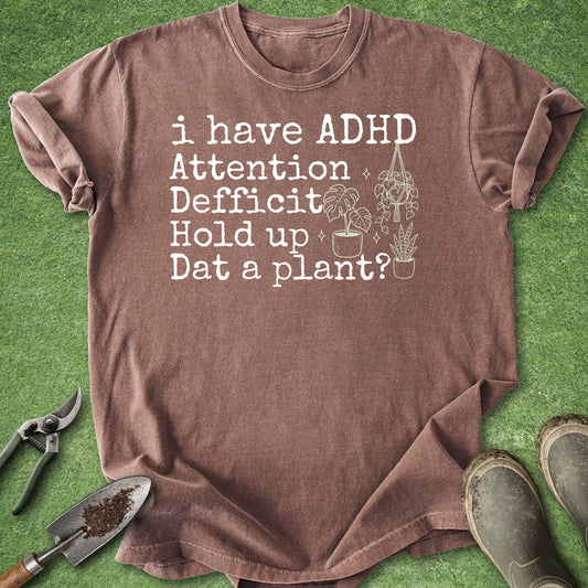 A brown short-sleeve t-shirt with a white graphic text reading 'i have ADHD Attention Deficit Hold up Dat a plant?' next to a small plant illustration, laid flat on green grass with gardening tools and boots around it.